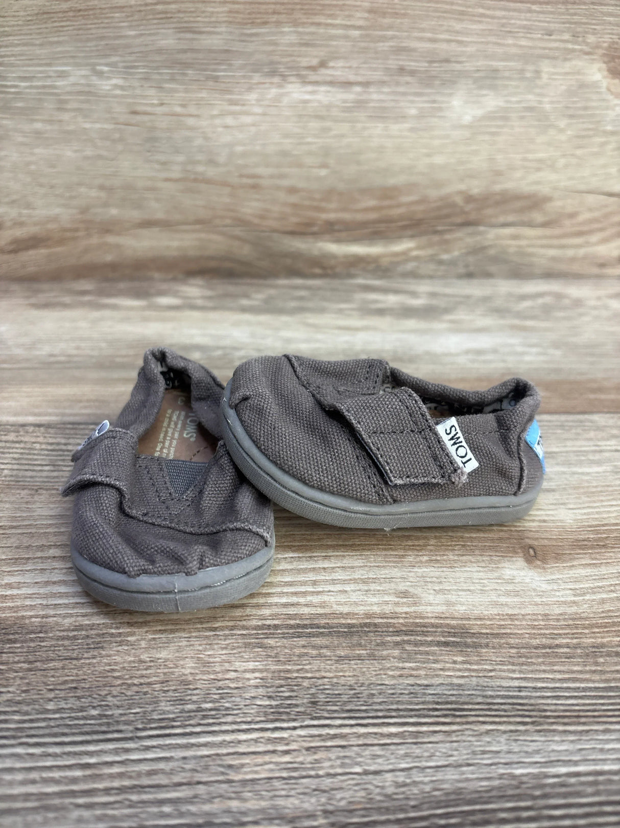 Angled front view of grey Toms Tiny Alpargata toddler slip-on showing canvas upper, Velcro strap and scuffed rubber sole on wood surface