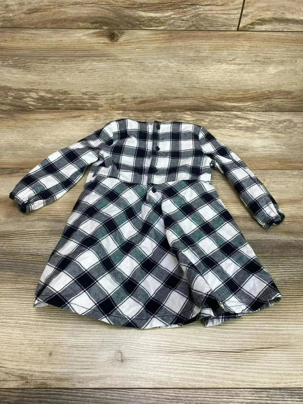 Flat-lay of white and black buffalo-check baby dress showing button-front bodice, long sleeves, and gathered skirt on wooden floor background