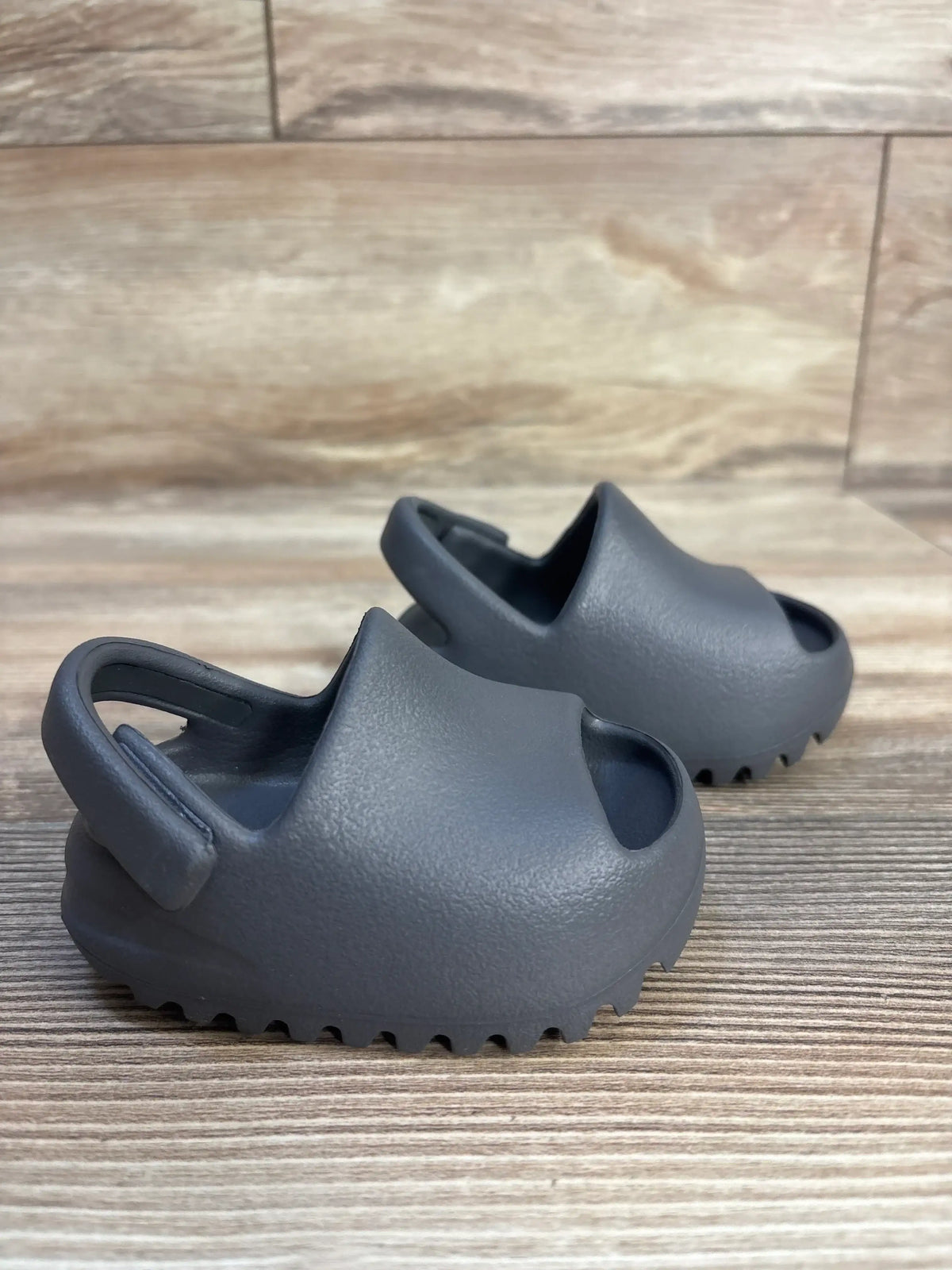 Three-quarter view of Adidas Yeezy Slides in Granit toddler size 3C showing closed-toe molded upper, back strap, and ribbed sole on wood floor