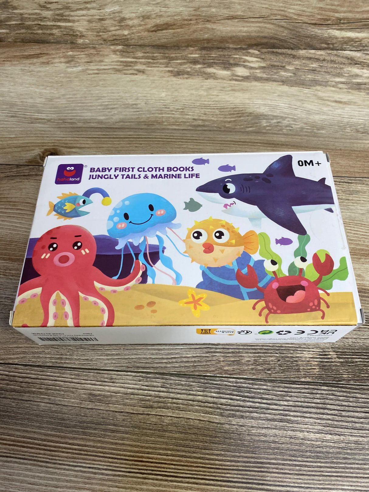 Angled top view of HahaLand Baby First cloth books 2-pack box showing headline Jungly Tails and Marine Life and colorful marine animal illustrations on a wood table
