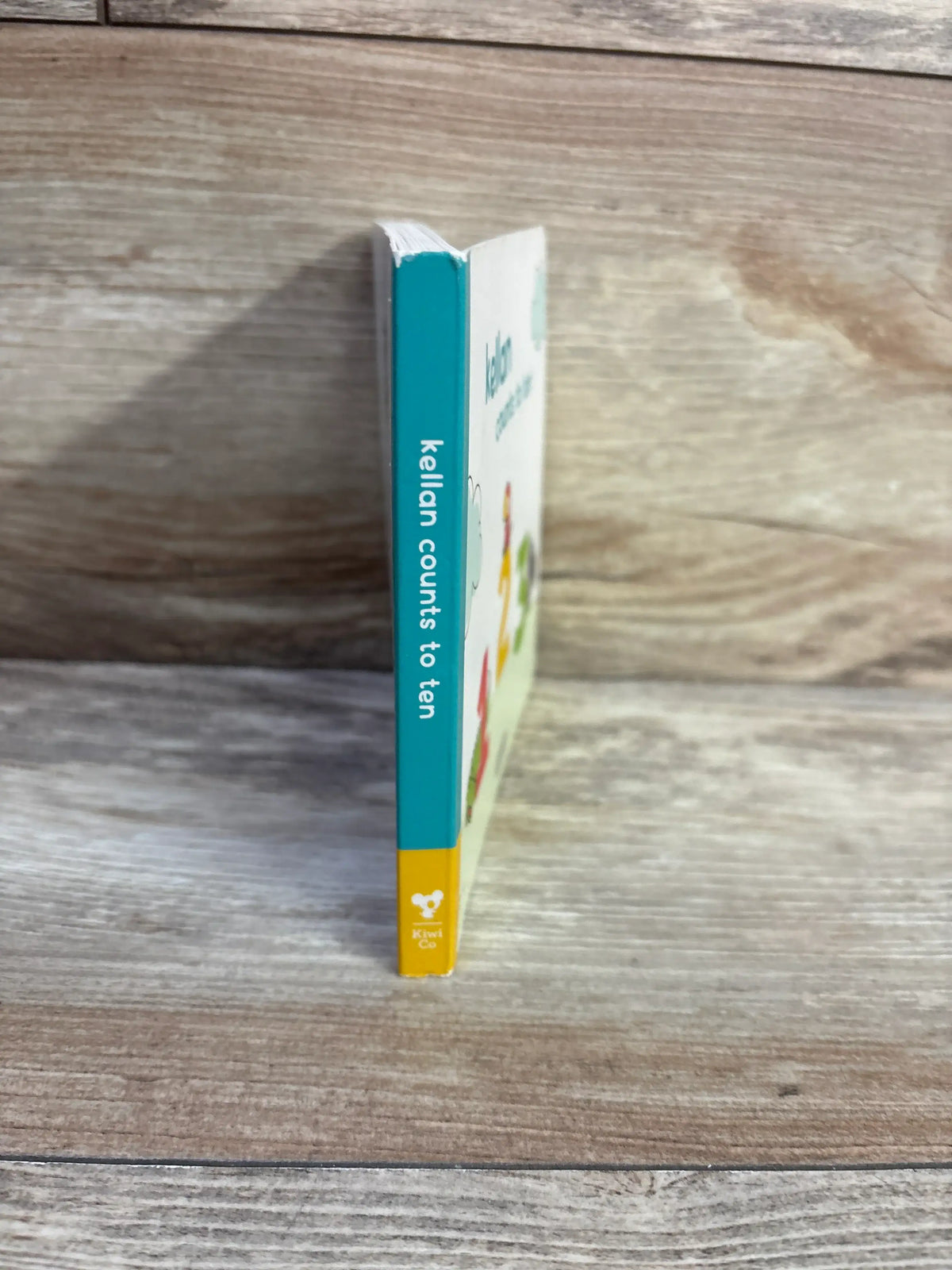 Side view of KiwiCo Kellan Counts To Ten board book showcasing its turquoise spine and yellow KiwiCo branding on a wood-grain surface