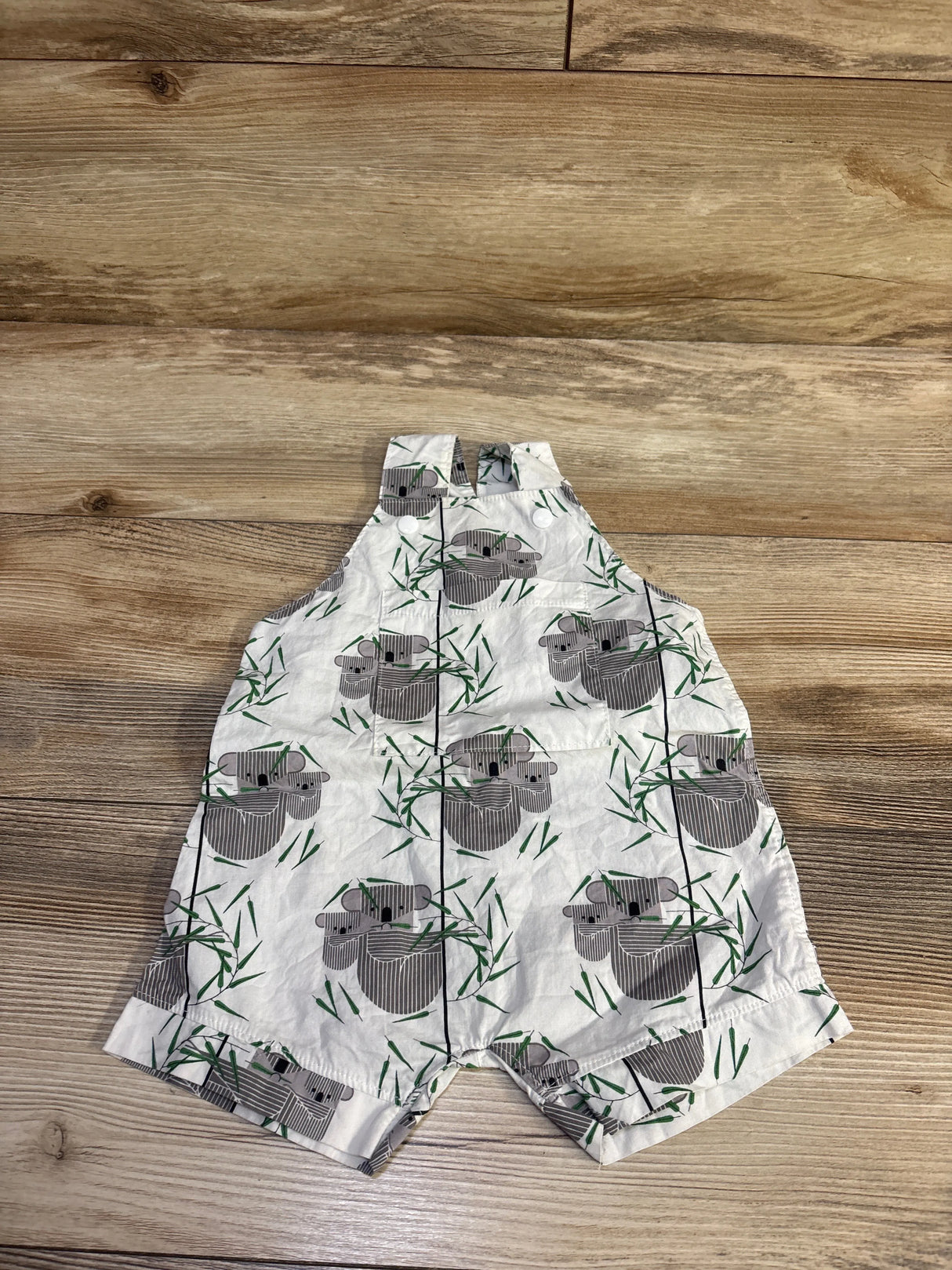 Flat lay of Little Stitches Taylor shortalls in white with koala and bamboo print, front button straps on wood background, size 6 months