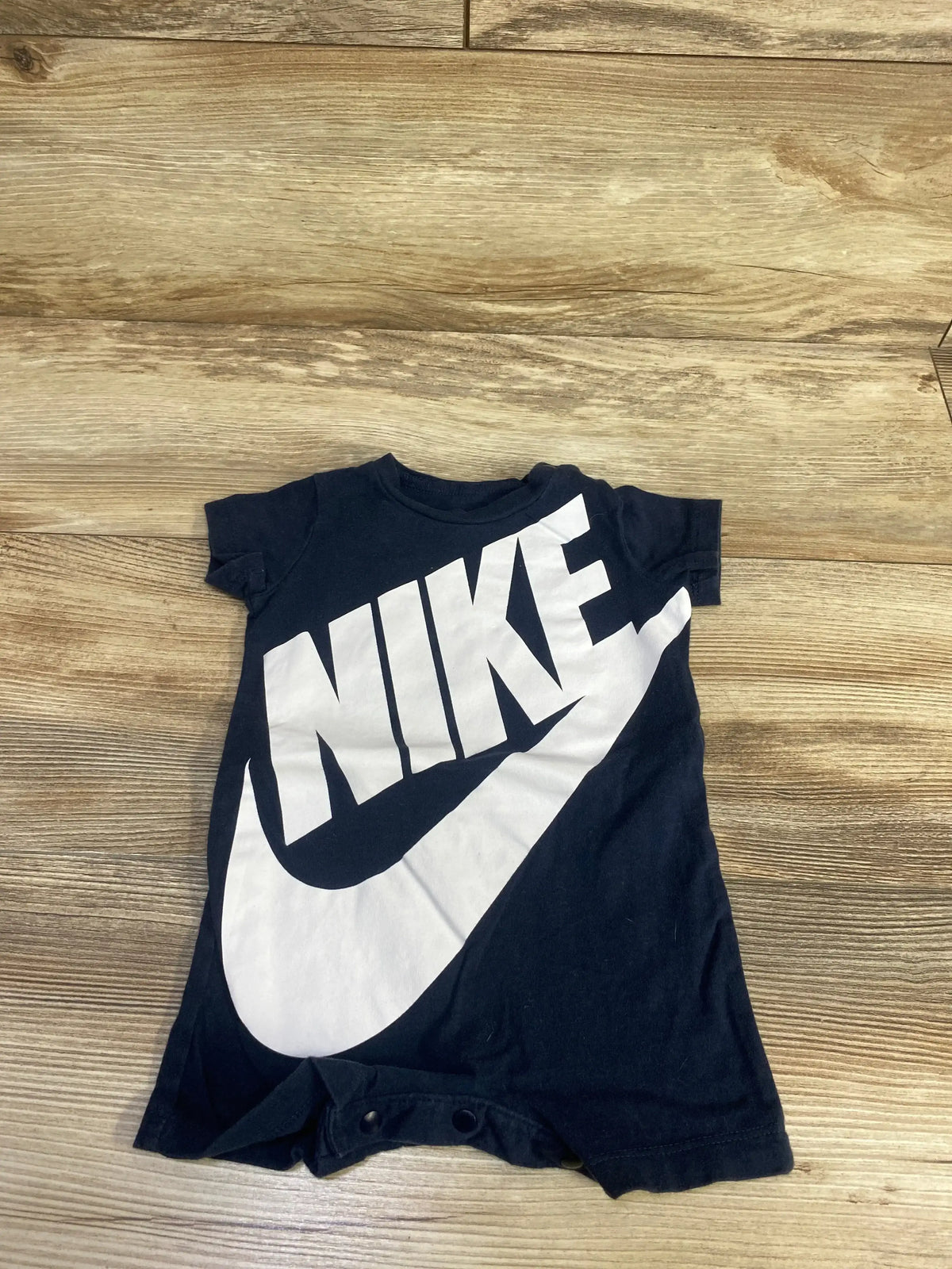 Front view of black baby Nike shortie romper showing large white NIKE logo and snap crotch closure on light wood floor