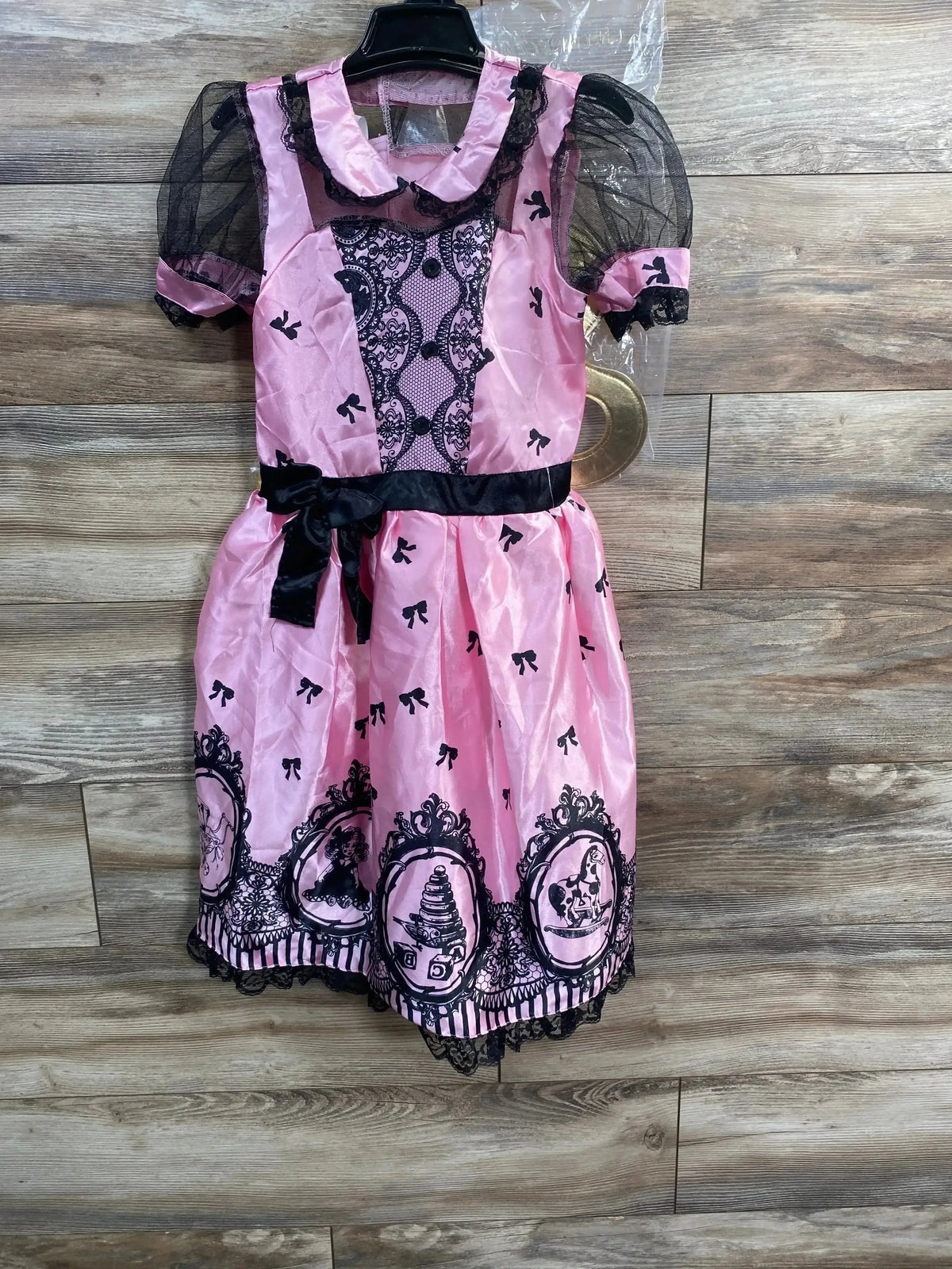 Front view of pink wind-up doll costume size 10-12 with black lace trim, puff mesh sleeves and black sash on wood background