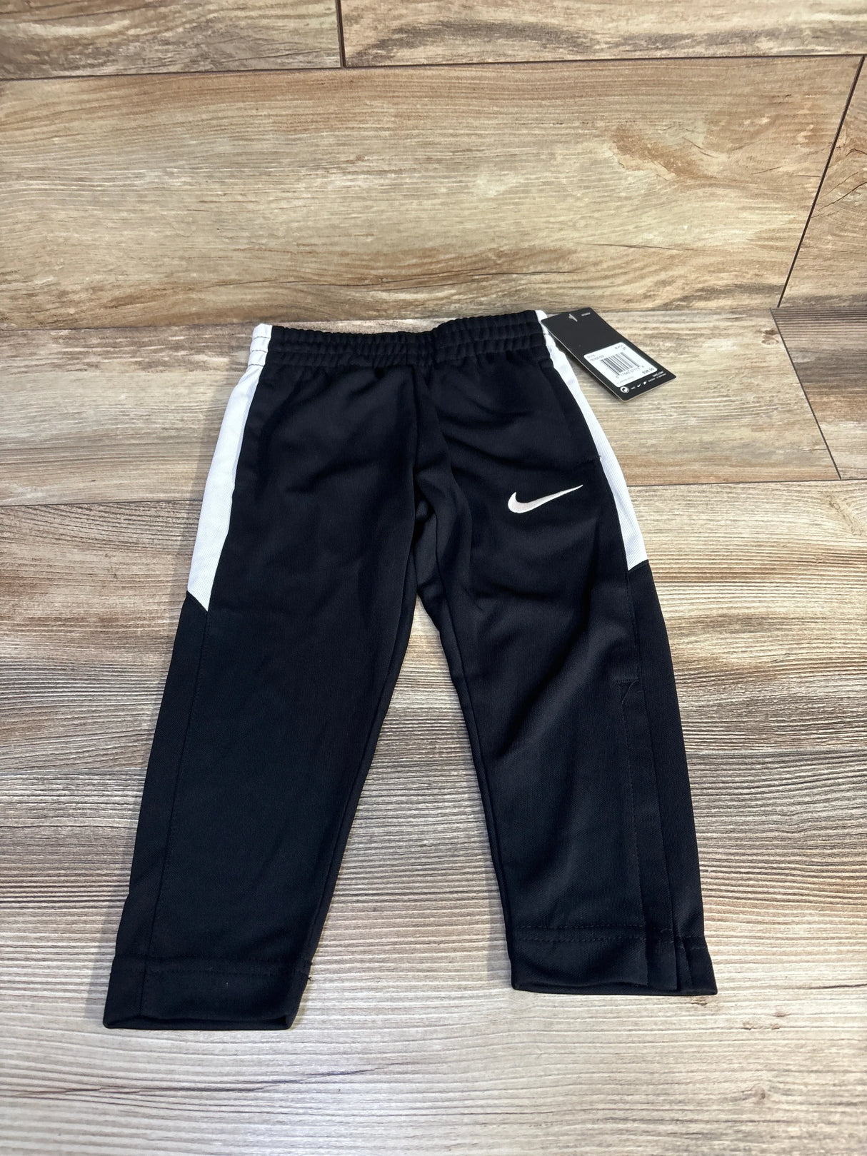 Front view of black Nike toddler joggers with white side panels, elastic waistband, and swoosh on wood background