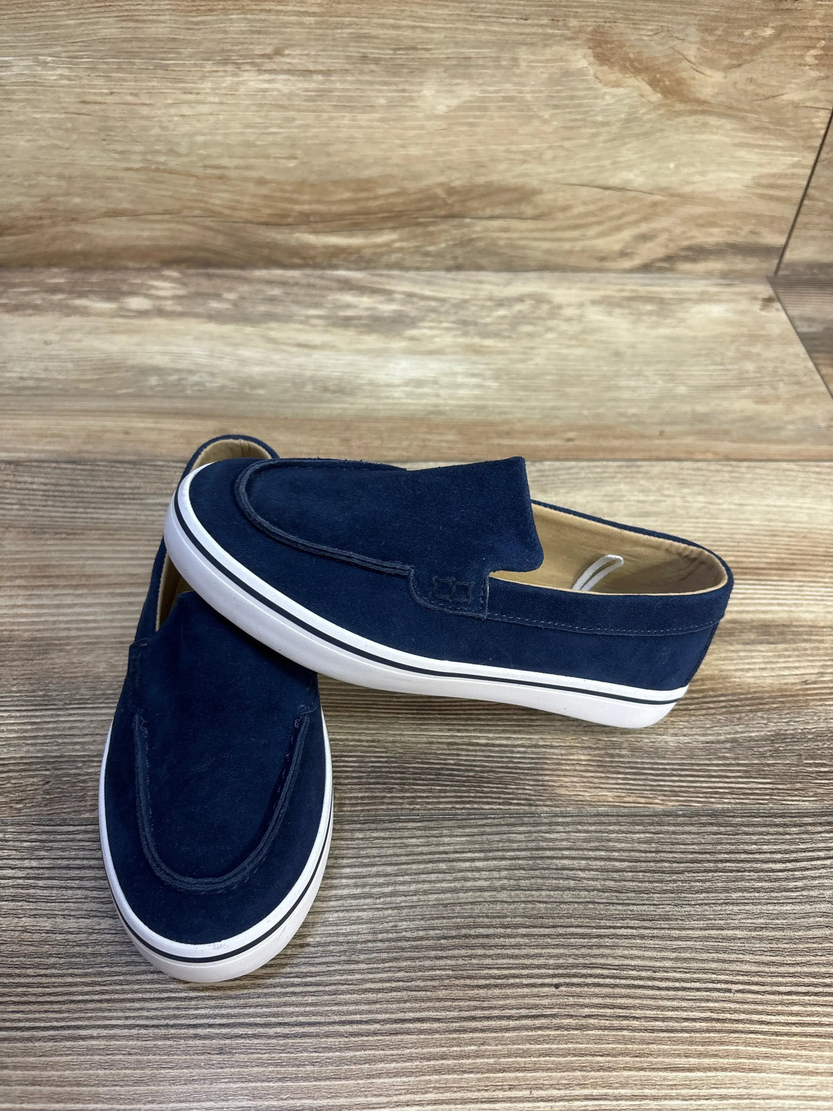 Angled view of Janie & Jack blue suede slip-on sneakers in size 12C showcasing white rubber soles on a wood-grain surface