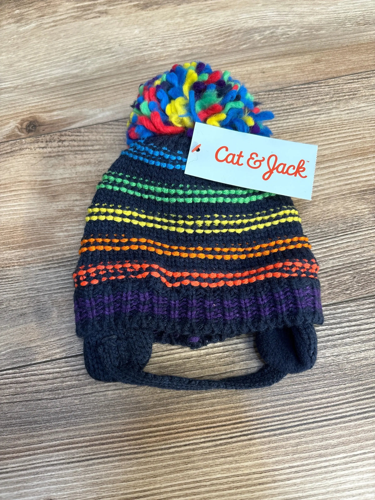 Kids knit beanie with rainbow stripes and pom-pom, ear flaps, on wooden background