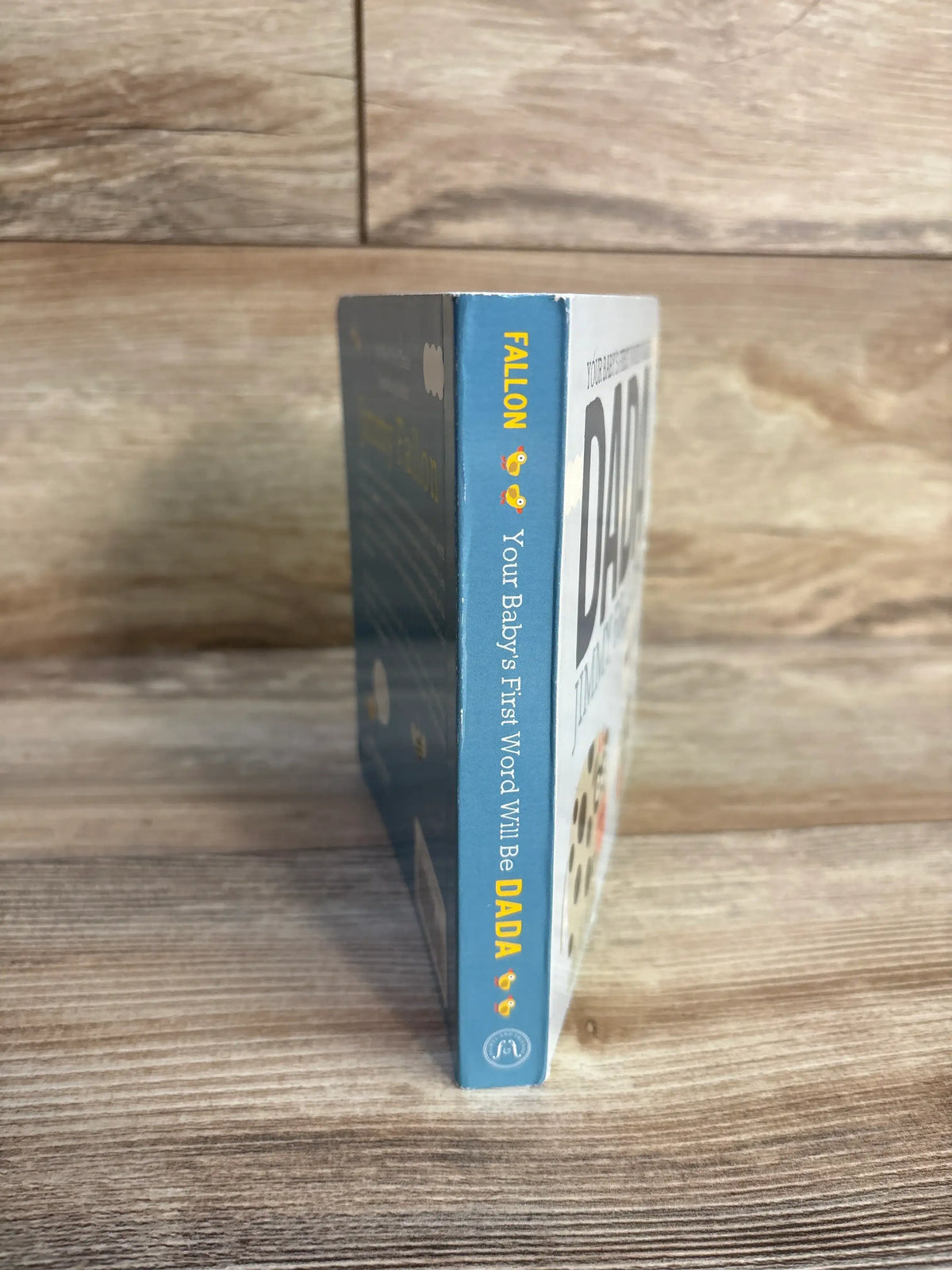 Side view of Your Baby’s First Word Will Be Dada board book by Jimmy Fallon showing its blue spine on a wood surface