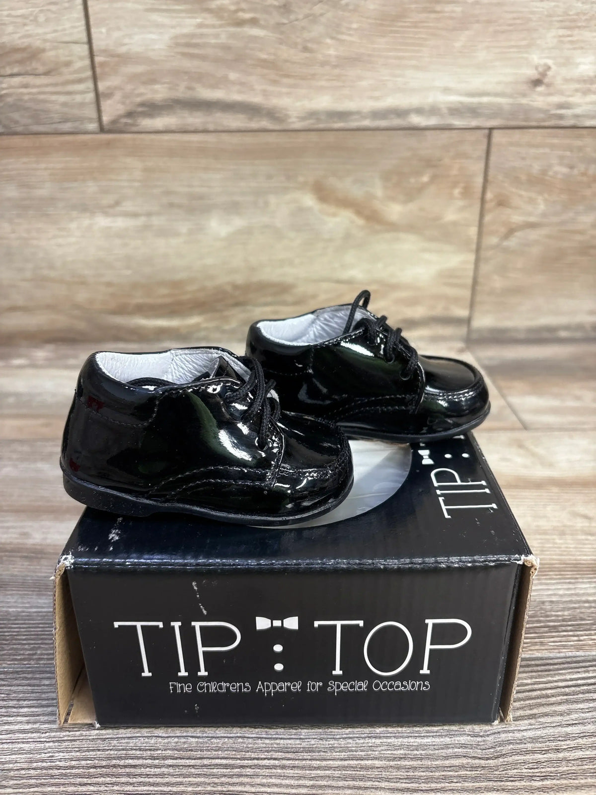 Side view of Tip Top genuine leather black baby sneakers showing glossy patent finish and laces on a branded box