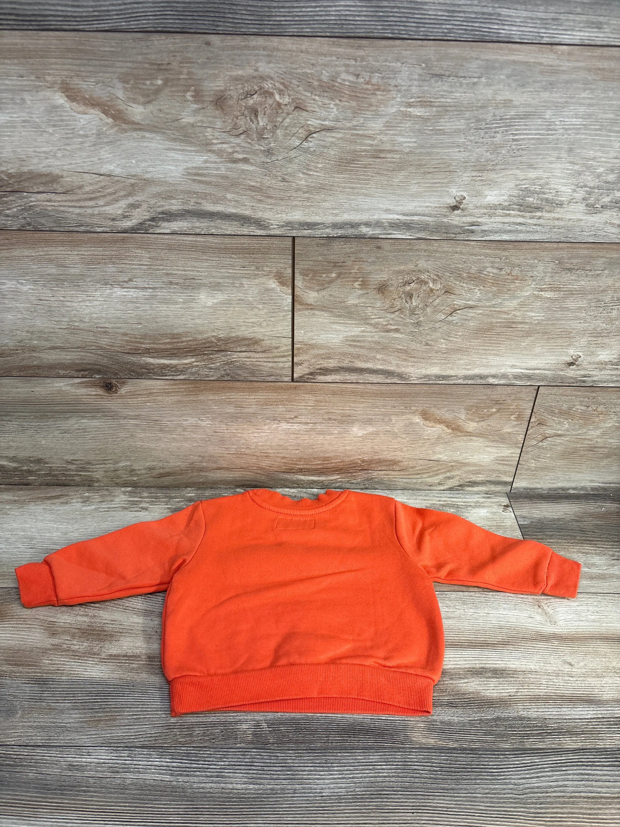 Back view of Old Navy Bord To Play Sweatshirt Blood Orange sz 12-18m showcasing solid orange crewneck with ribbed cuffs and hem on wood plank floor