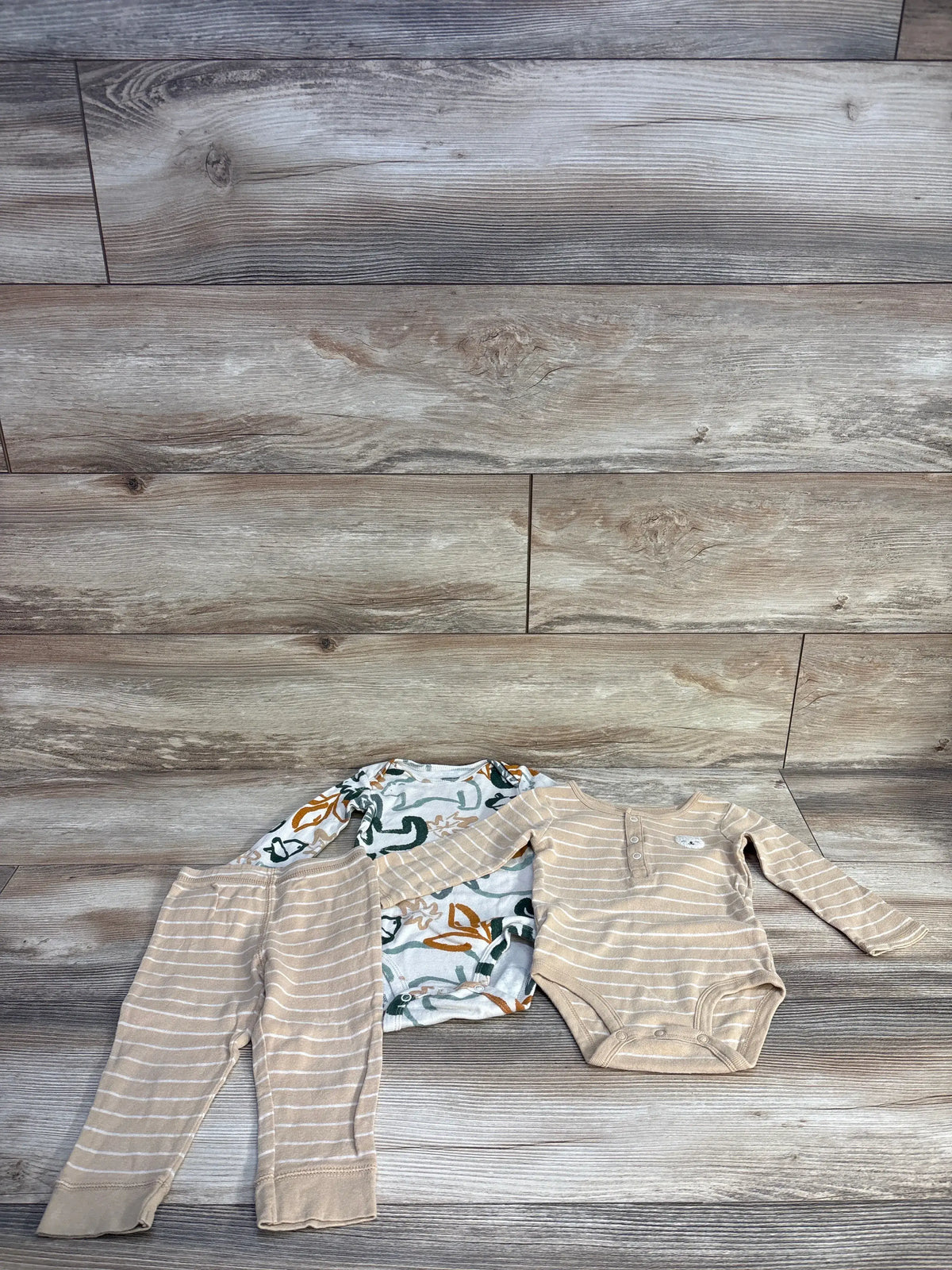 Top view of Carter’s three-piece beige bodysuit set with striped pants, patterned bodysuit, and snap closures on wood flooring