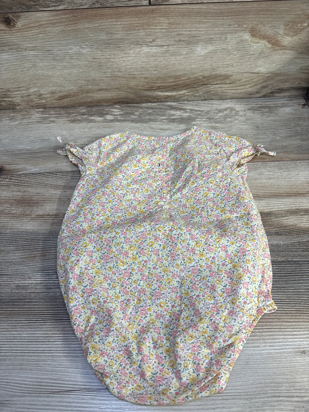 Flat lay of yellow OshKosh floral bubble romper (18m) showing cap sleeves with ties and elastic leg openings on wood background