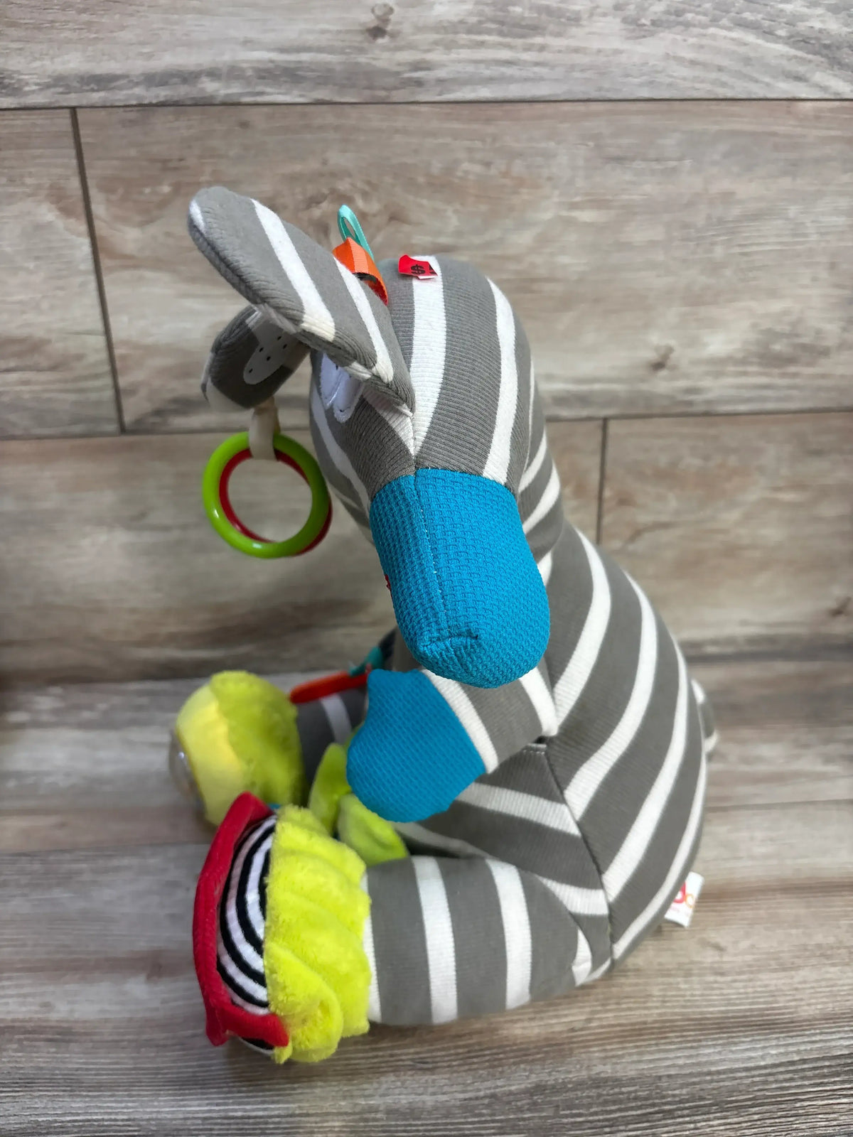 Side view of Dolce Zeddy The Zebra Activity Plush showcasing gray and white stripes, blue accents, and activity attachments on a wood floor