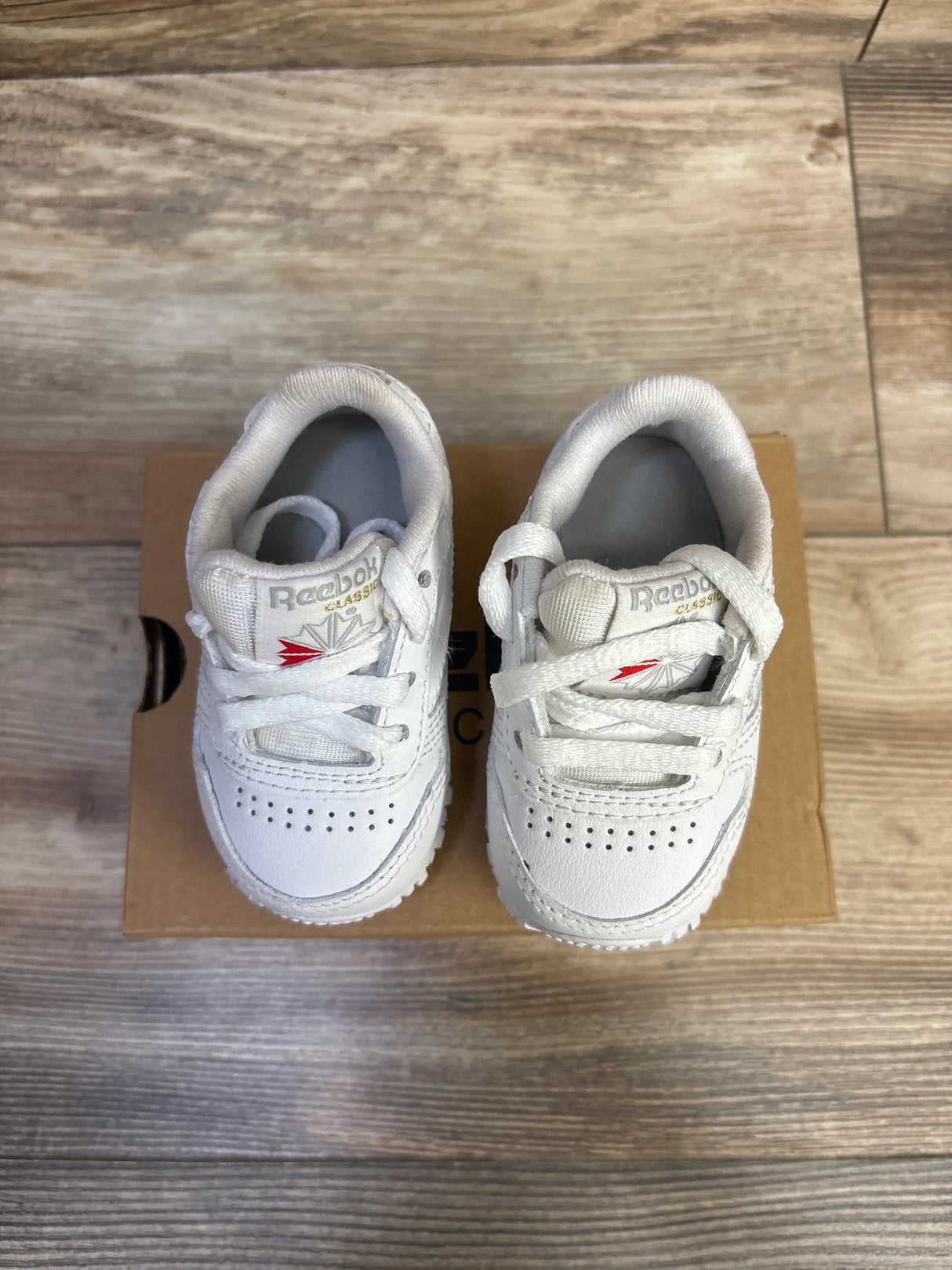 Top view of white Reebok infant sneakers showing perforated toe, laces, and red logo on a cardboard box on wood floor
