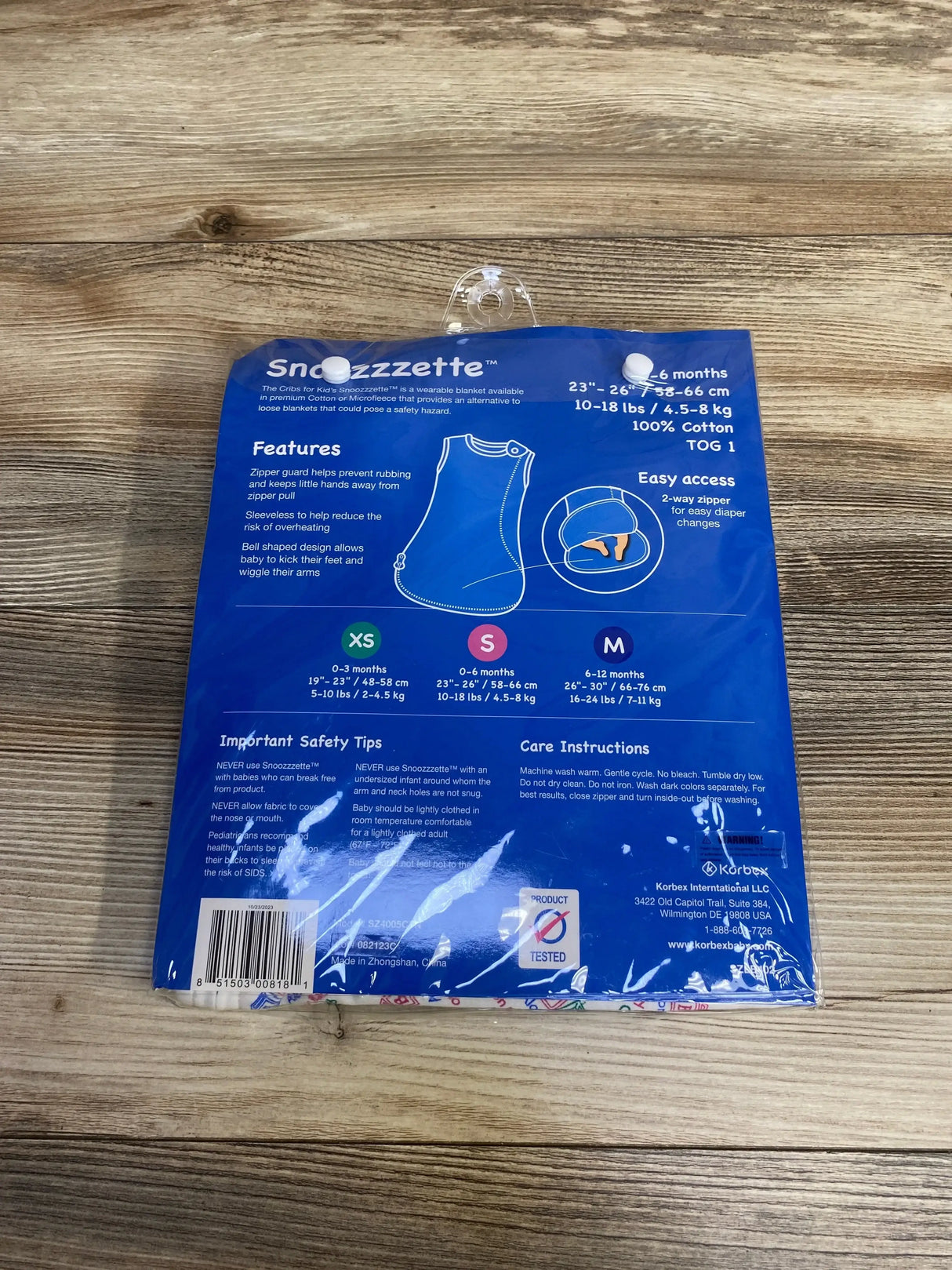 Back of packaged Cribs for Kids Snoozzzette wearable blanket showing size chart and features on blue card over wood surface