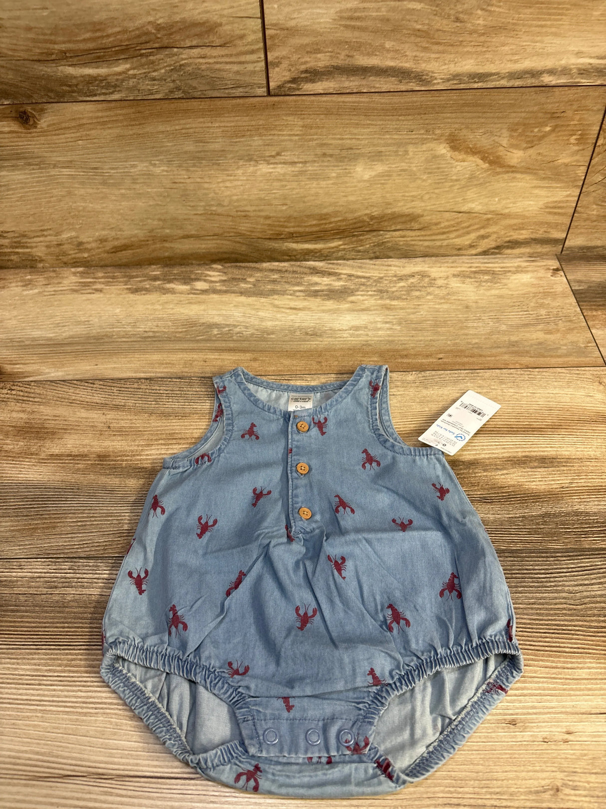 Top-down view of Child of Mine chambray henley bubble romper with three wooden front buttons and red lobster print on light-blue fabric, on wood background