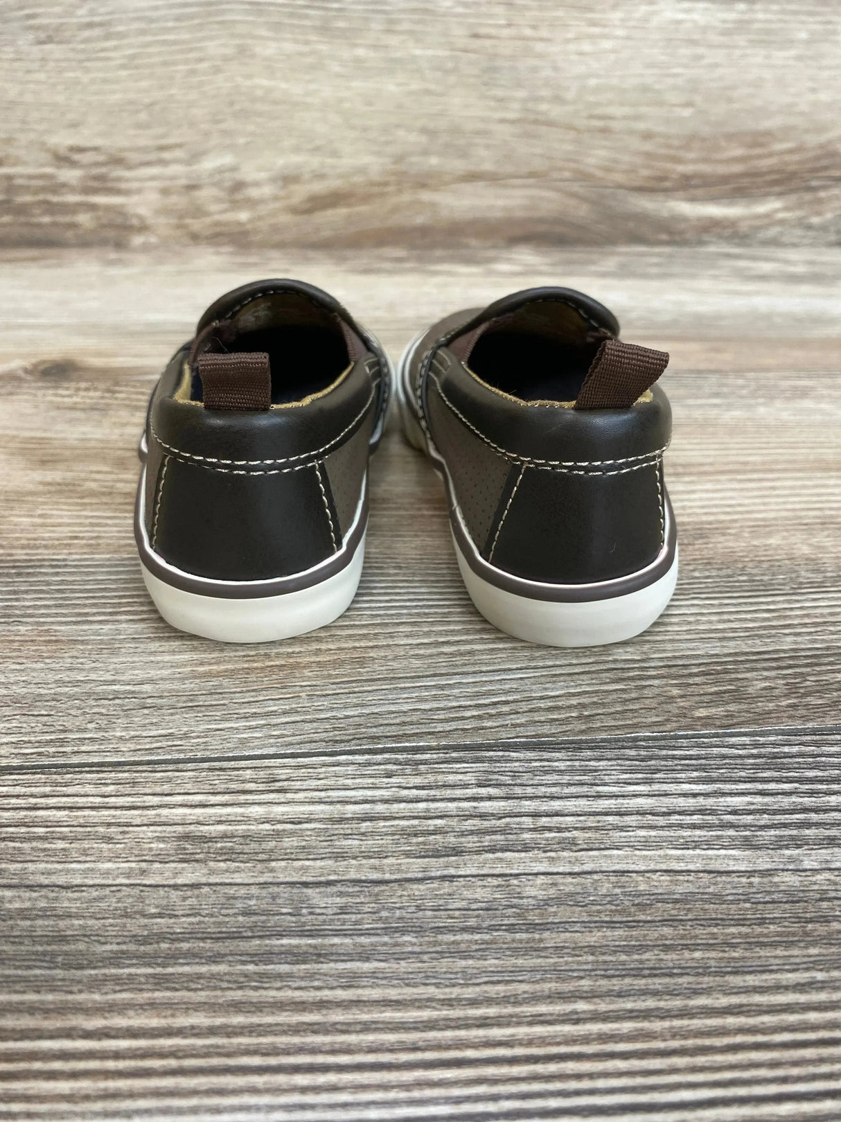Rear view of brown toddler slip-on sneakers with white soles, contrast stitching and heel pull tabs on wood background