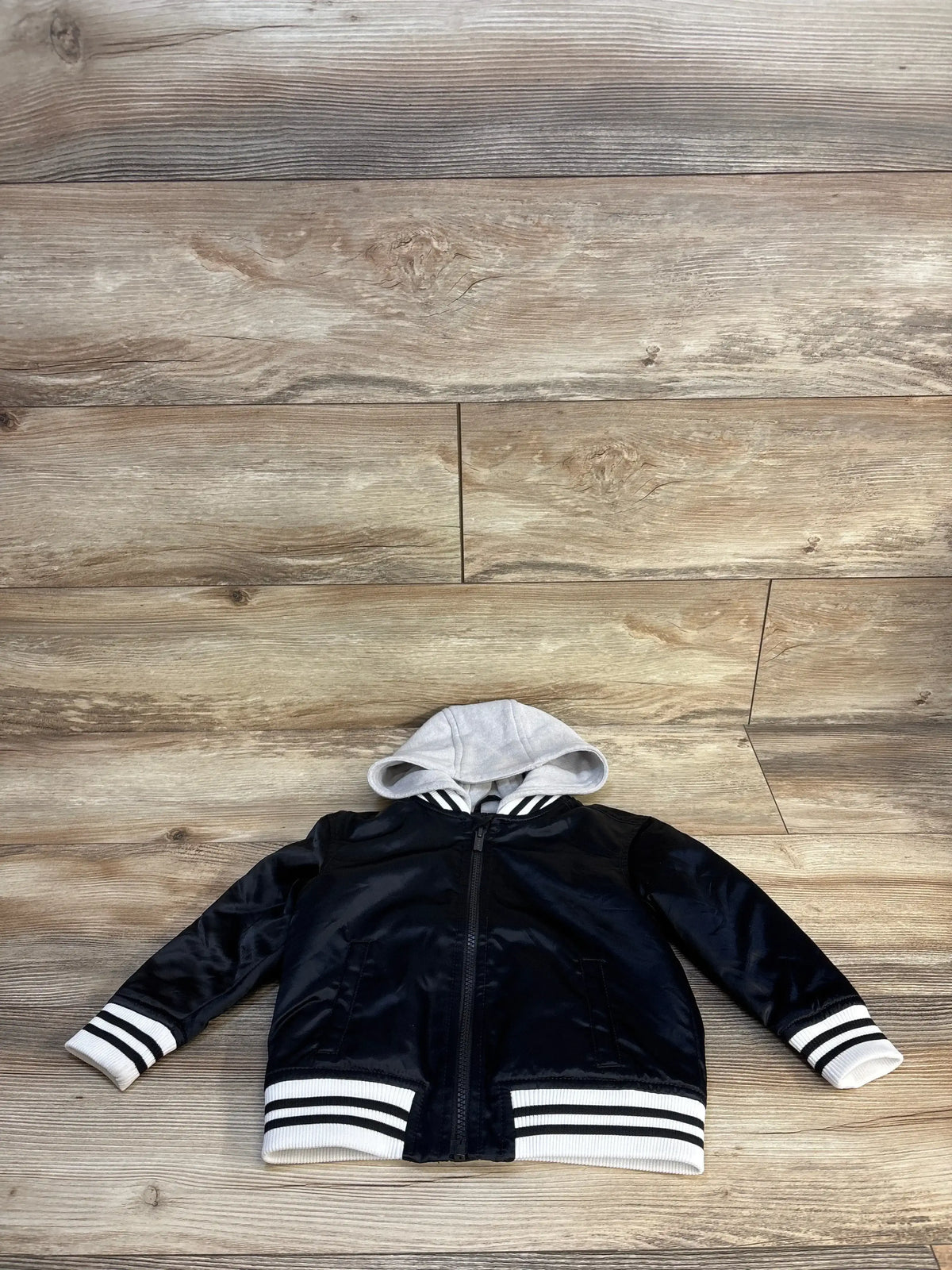 Black kids bomber jacket with gray hood and striped cuffs, laid on wood floor
