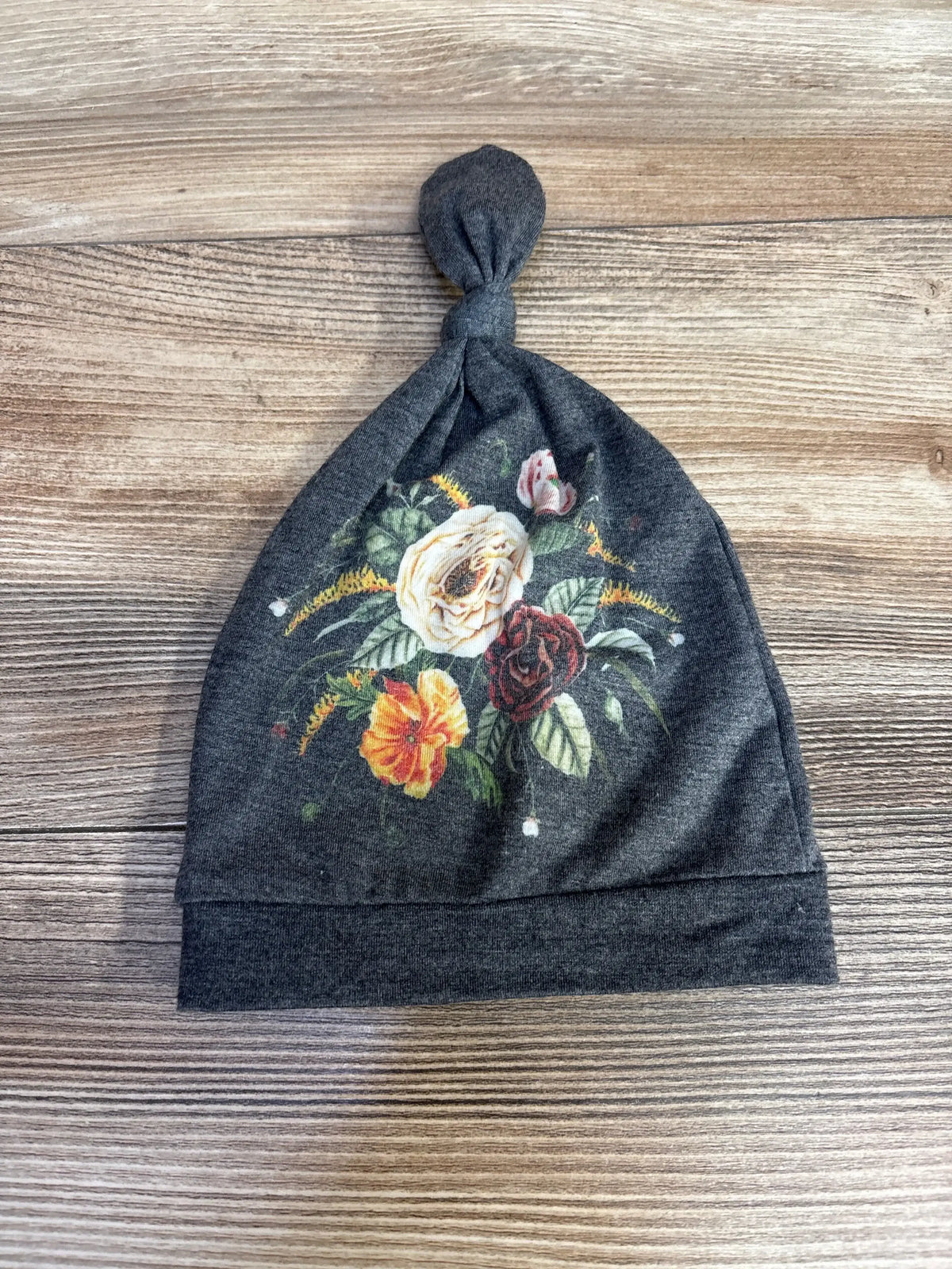 Flat lay of Copper Pearl floral top knot baby hat in grey showcasing knotted top and multicolor floral print on wood background