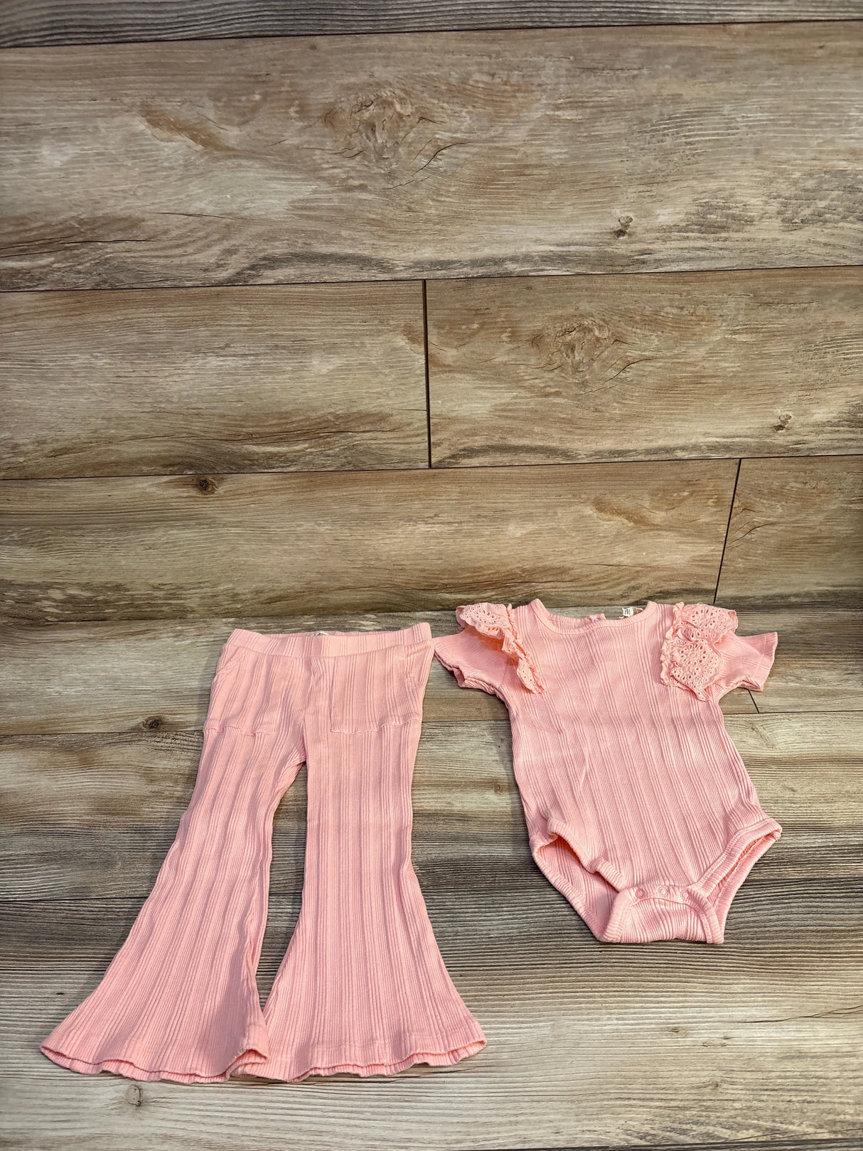 Flat lay of pink ribbed two piece baby set with eyelet trim ruffle sleeve bodysuit and matching flared pants on wood