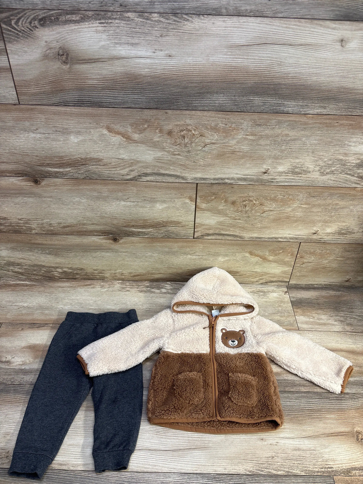 Flat lay of Little Me faux-fur jacket and pants set hooded beige-brown bear jacket with pockets and dark gray pants on wood