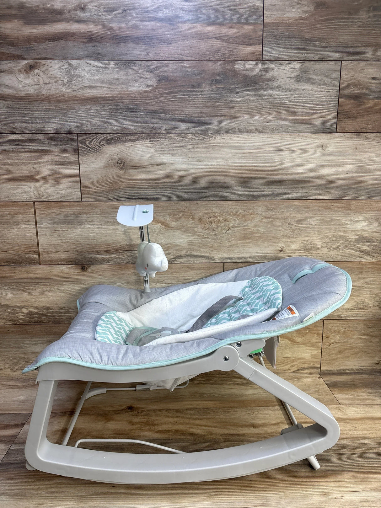 Side view of Ingenuity Keep Cozy 3-in-1 bouncer showing padded grey and mint seat, toy bar, and plastic rocker on wood floor