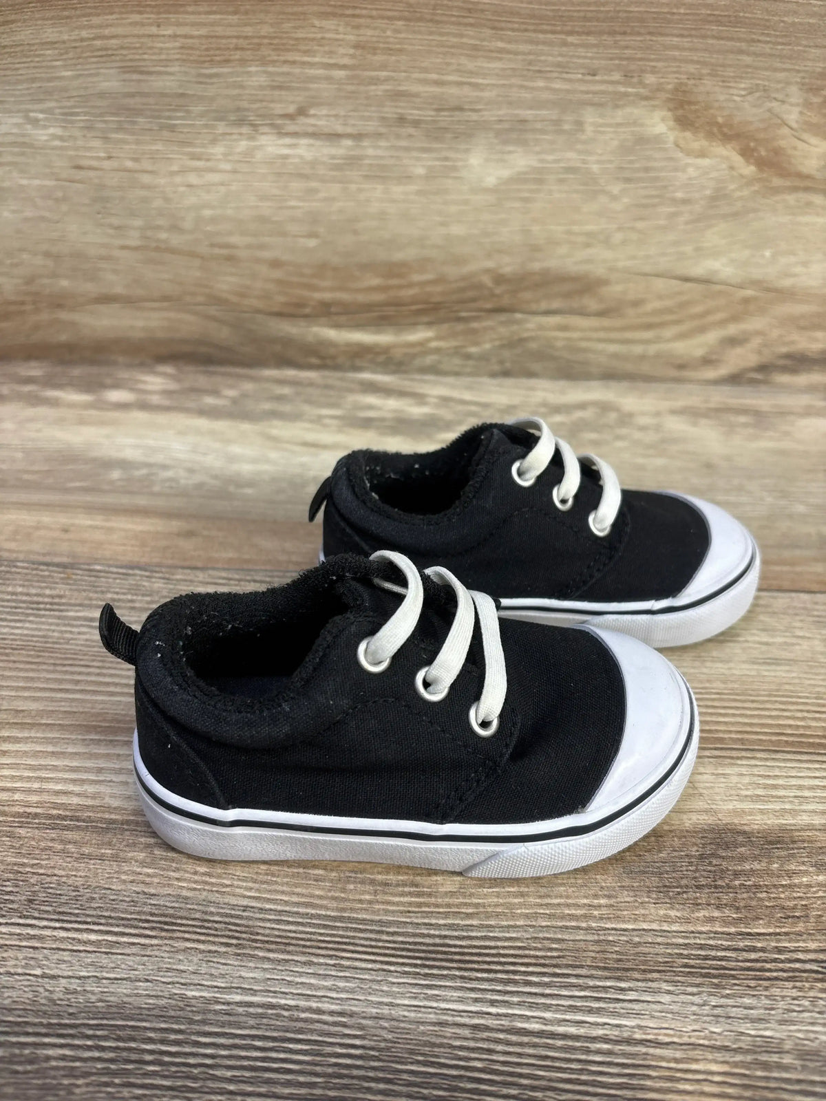 Side view of toddler black canvas bump-toe sneakers showcasing white rubber toe cap and laces, size 5C on wood background