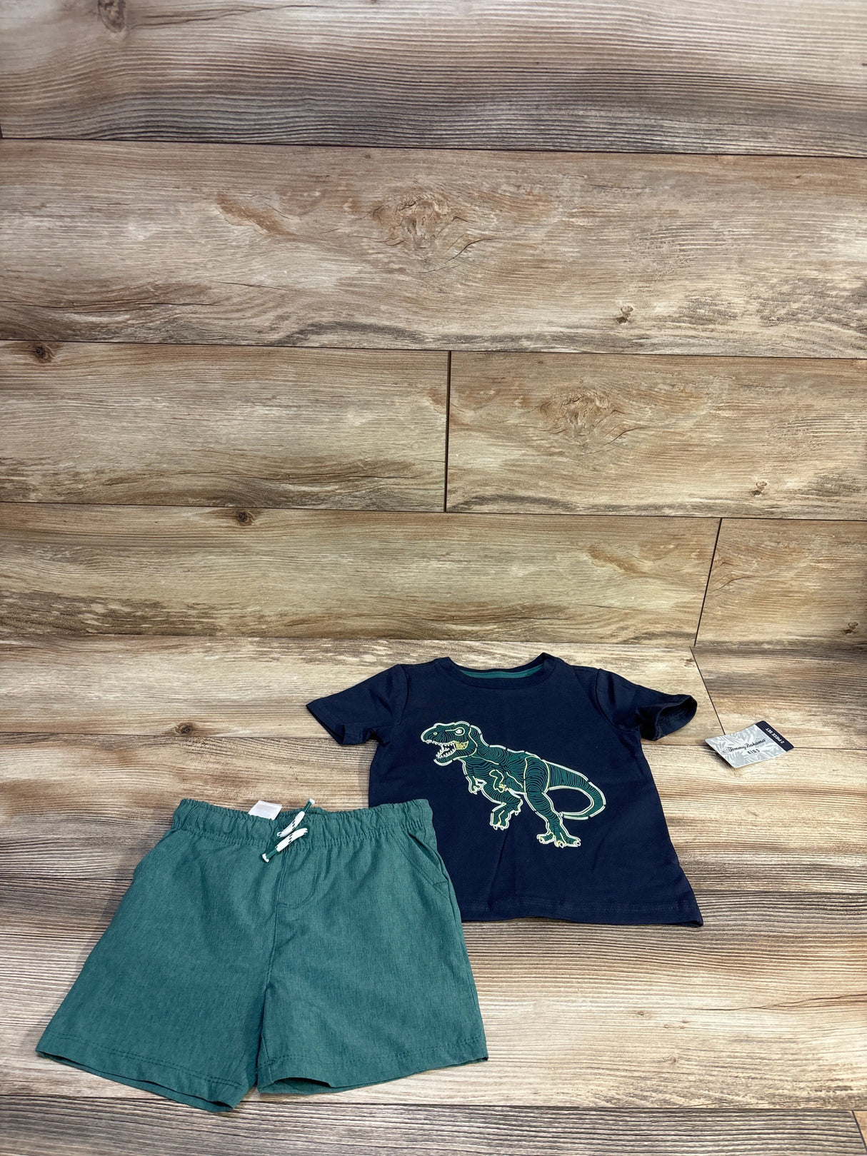 Flat lay of toddler navy Tommy Bahama T-shirt with green dinosaur graphic and teal board shorts on wood background