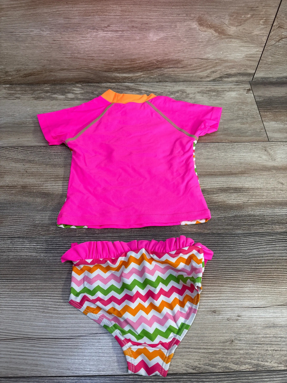 Flat-lay of girls’ 2-piece rashguard set showcasing bright pink short-sleeve rashguard with orange collar and chevron-print bikini bottoms with ruffle waist on light wood background