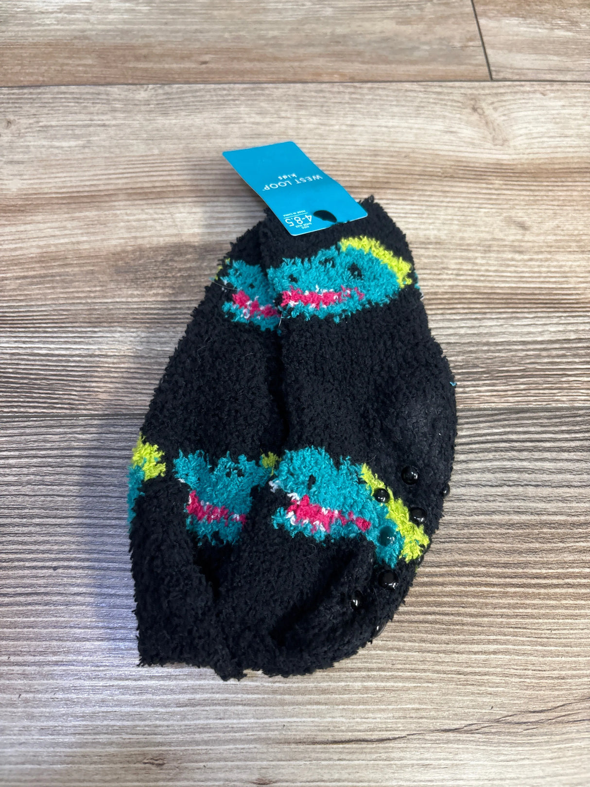 Top-down view of black West Loop kids fuzzy socks size 4-8.5C showcasing teal dinosaur pattern and black gripper dots with wood floor background