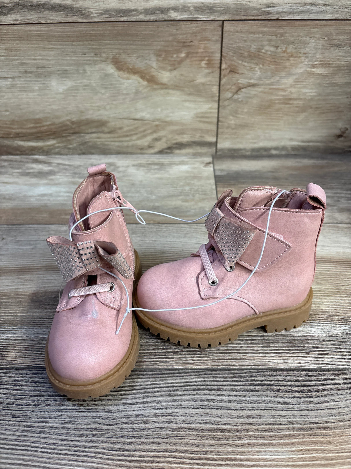 Front three-quarter view of Jessica Simpson Daria pink toddler ankle boots showcasing rhinestone bow strap and tan lug sole on wood background