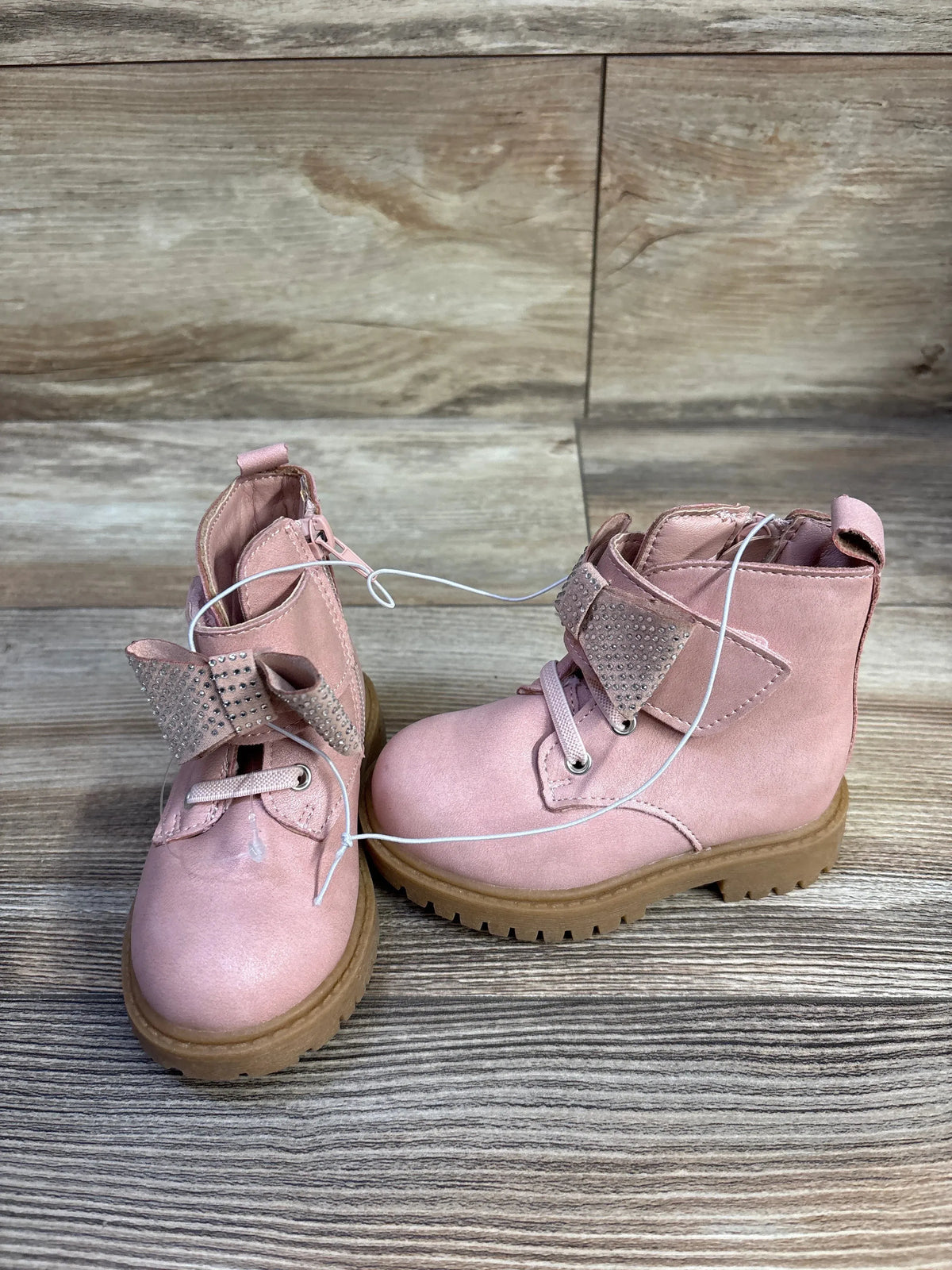 Front three-quarter view of Jessica Simpson Daria pink toddler ankle boots showcasing rhinestone bow strap and tan lug sole on wood background