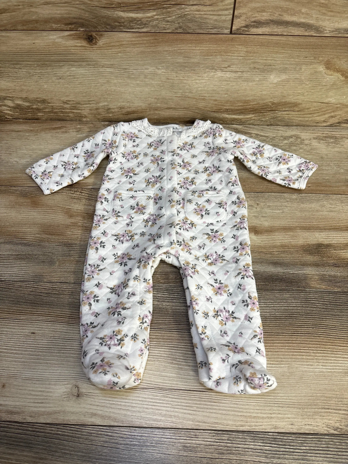 Front view of Nicole Miller white floral quilted footed coverall showcasing long sleeves and pockets on a wood floor