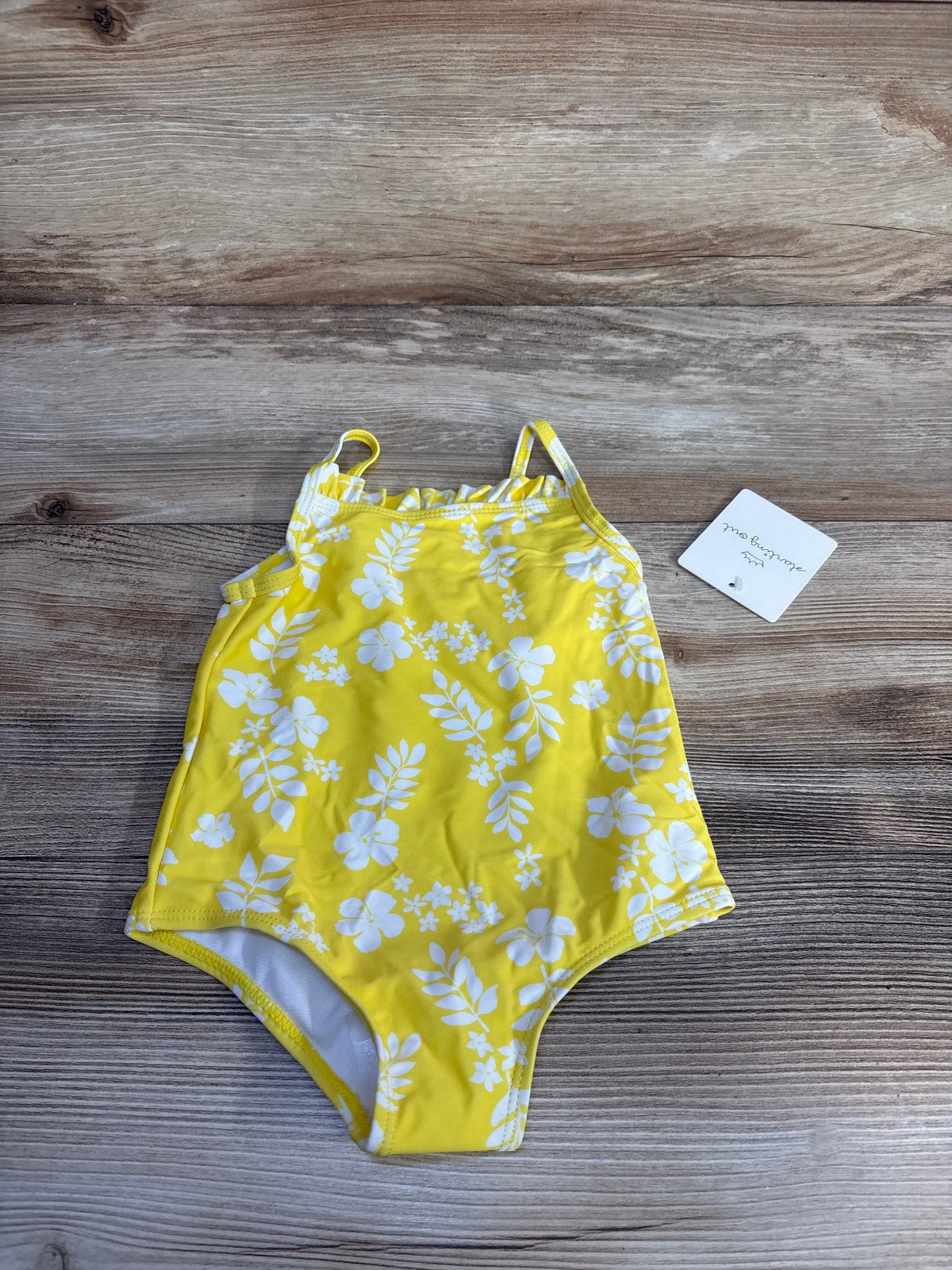 Front view of a yellow Starting Out one-piece swimsuit showcasing its white floral pattern on a wood-grain background