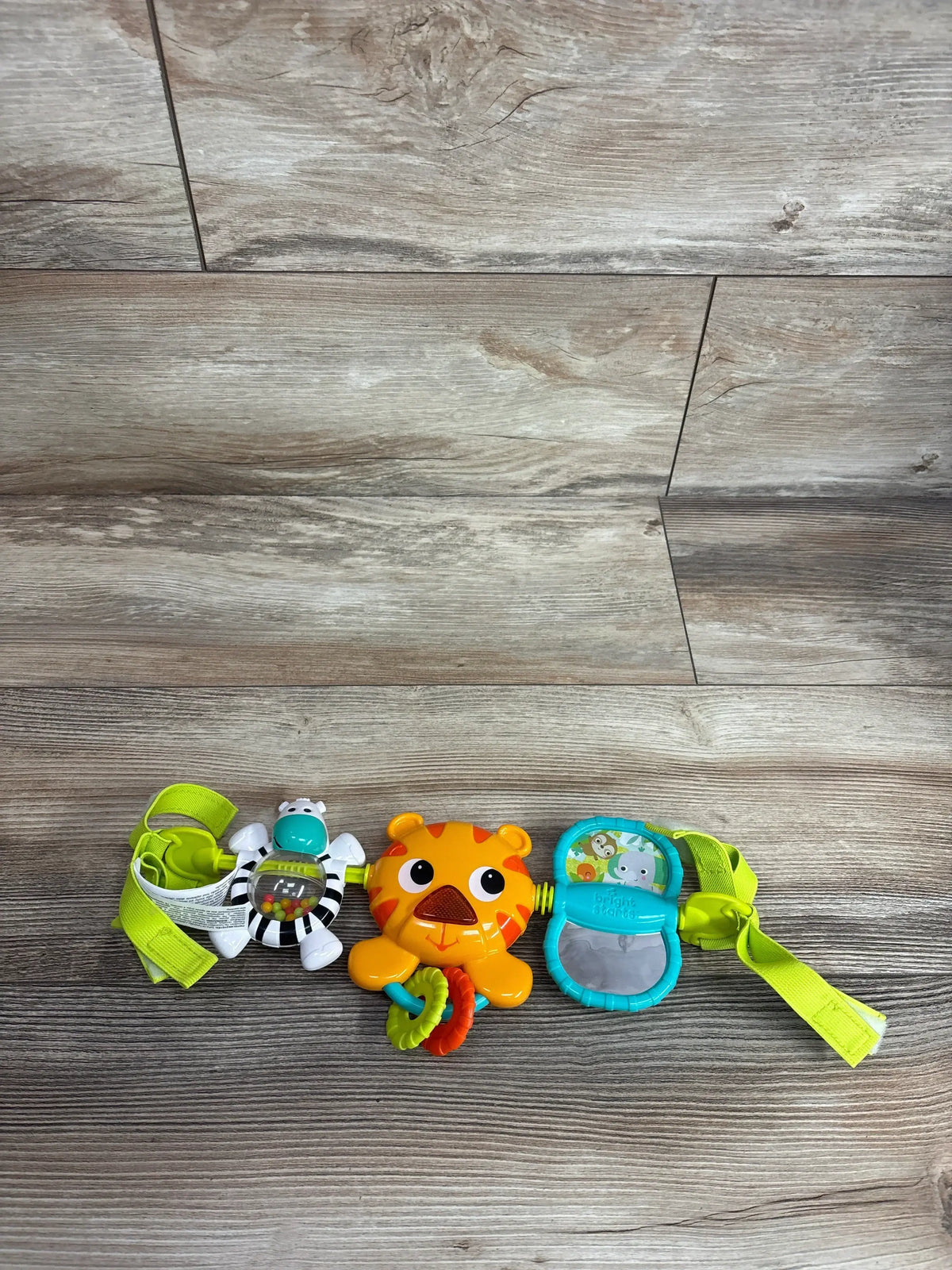 Top view of Bright Starts Take Along Carrier Toy Bar showcasing green strap with zebra tiger and mirror dangling toys on wood plank background