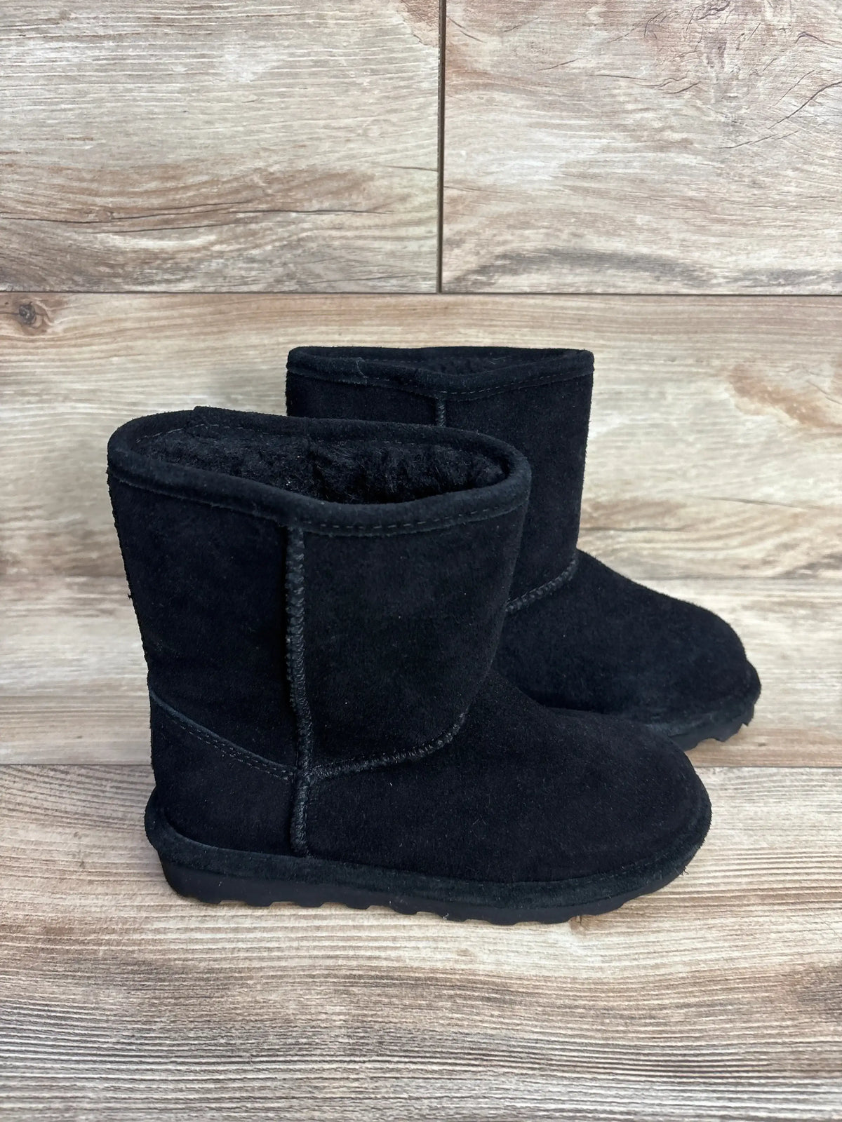 Three-quarter view of toddler Bearpaw Elle boots showing black suede, plush lining, and treaded sole on wood background