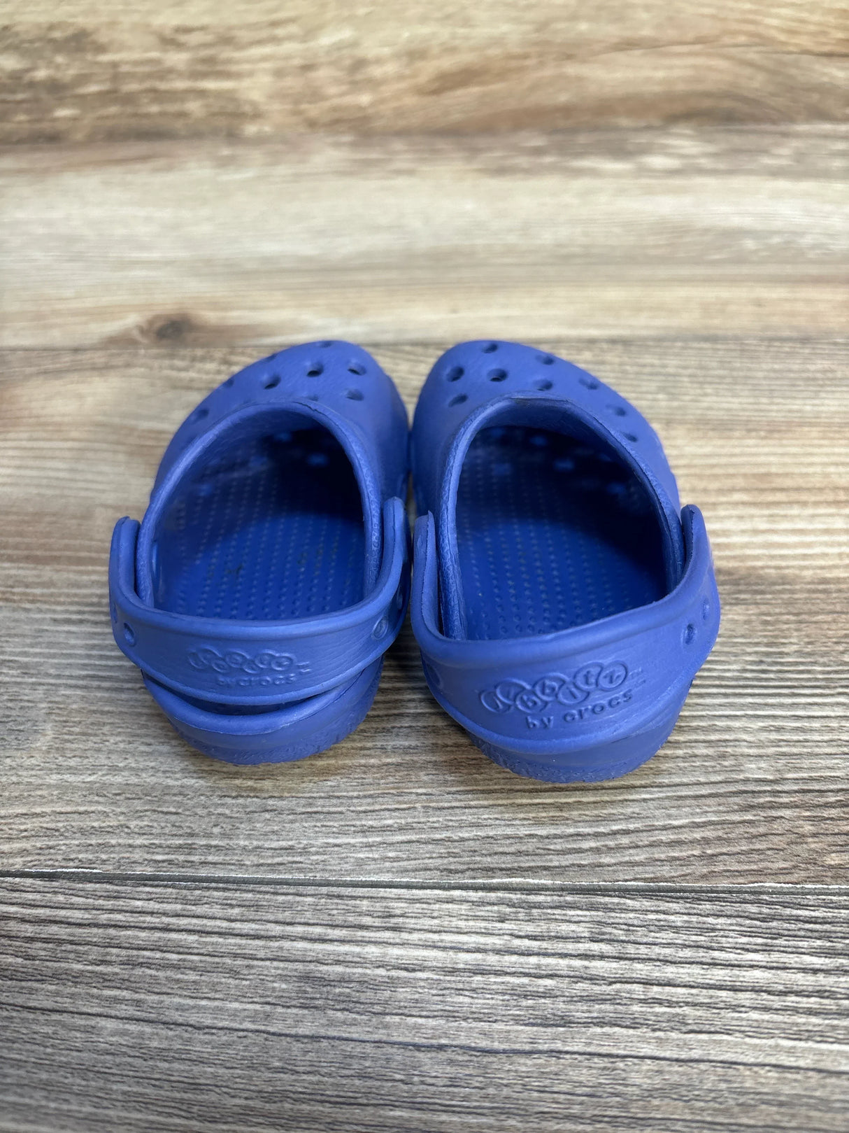 Rear view of toddler Crocs Classic clogs in blue showcasing adjustable back straps and ventilation holes on a wood floor