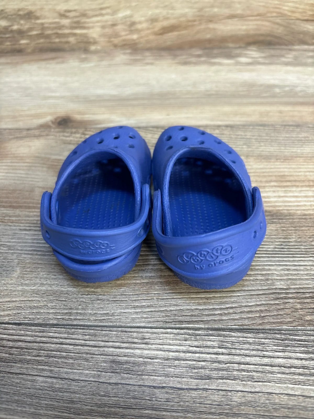 Rear view of toddler Crocs Classic clogs in blue showcasing adjustable back straps and ventilation holes on a wood floor