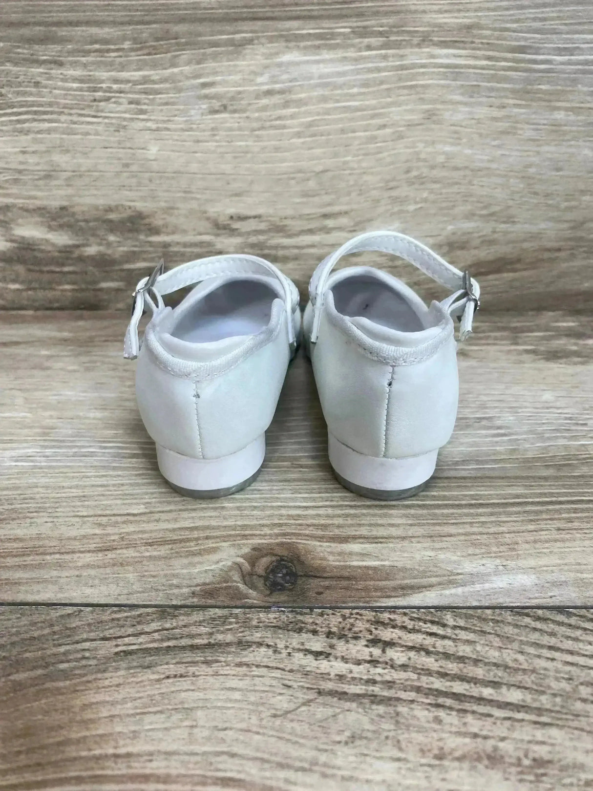 Rear view of white Theatricals tap buckle toddler shoes showing ankle straps with buckles and low heels on wood floor background