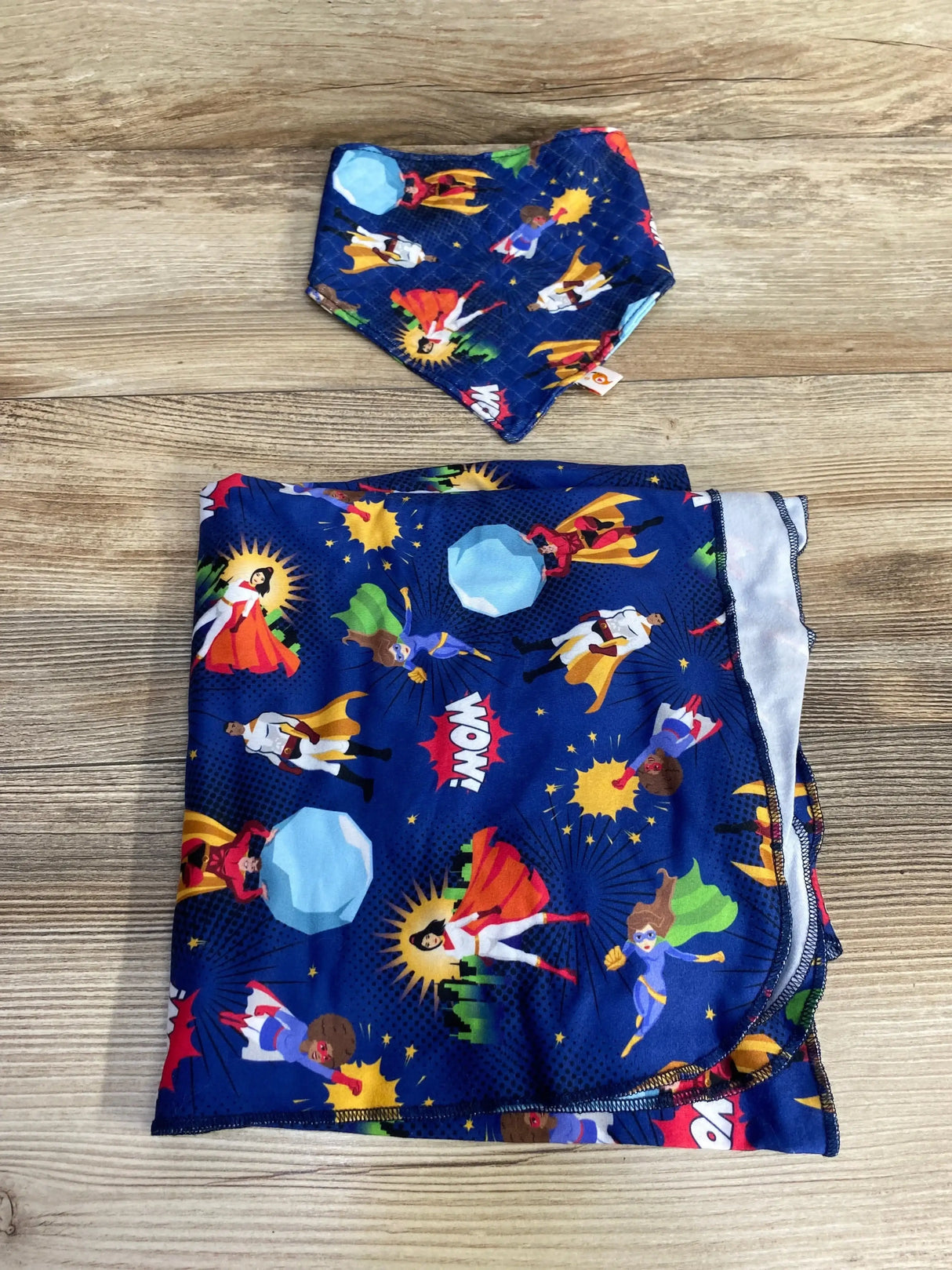 Folded Bumblito stretch swaddle set showing blue Never Alone superhero print on wood background
