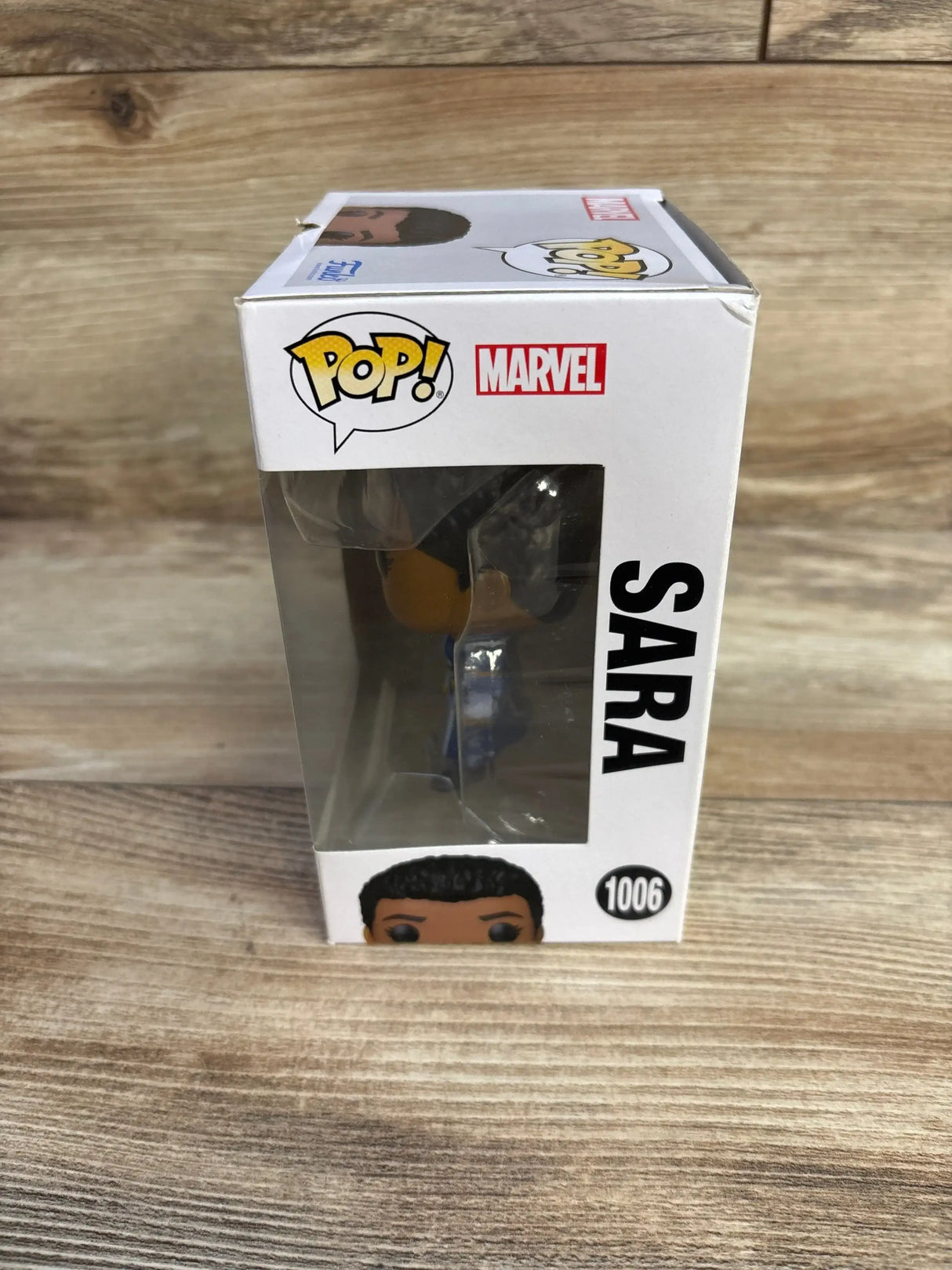Front-right view of Funko Pop Sara vinyl figure in original box showing clear window, SARA side text and Marvel and POP logos on wood plank background