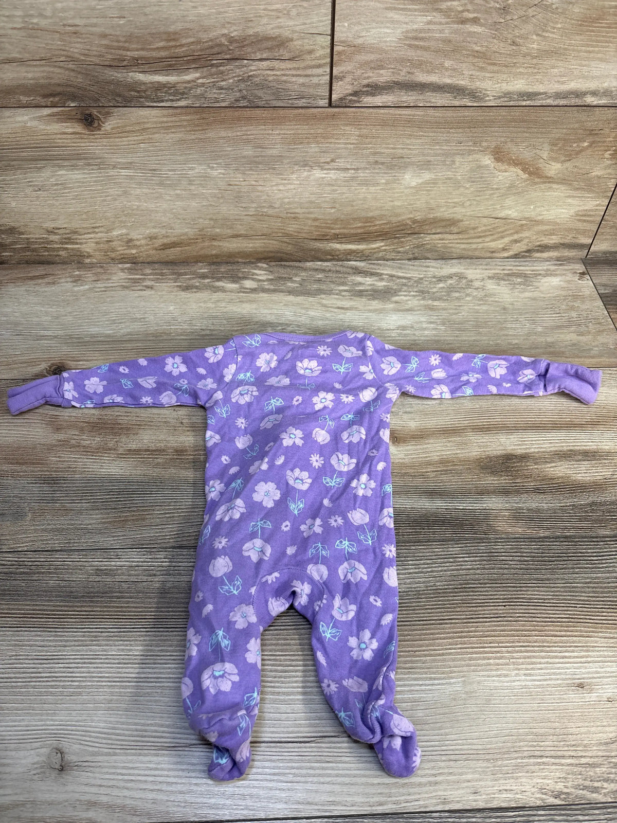 Top-down view of Member’s Mark floral sleeper in purple, showcasing long sleeves and footed legs on a wood-look floor