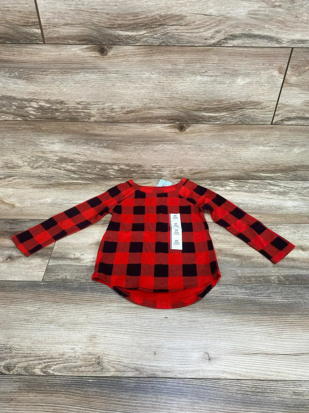 Flat lay of toddler red and black buffalo check long-sleeve shirt, size 2T, showing curved hem and crew neckline on wood floor background