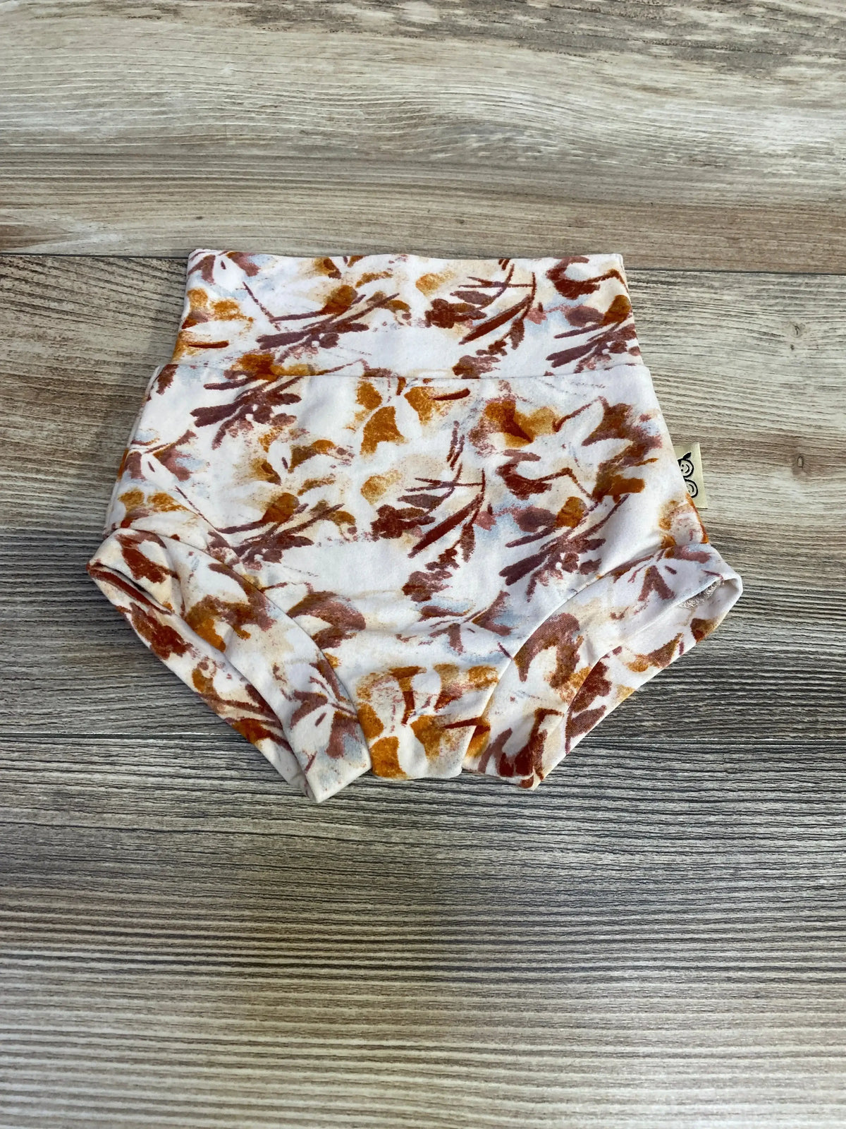 Top-down view of beige baby bummie diaper cover size 3-6m showcasing rust and mustard floral print on wood background