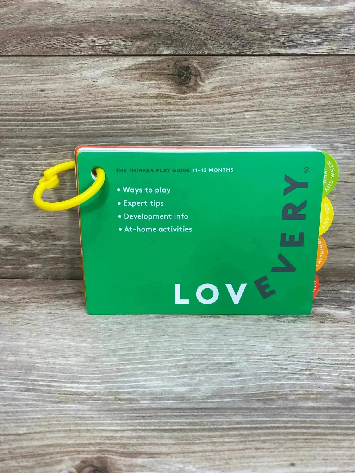 Front view of Lovevery 11-12 months play guide showing green cover, bullet list of tips, and yellow ring on wood background