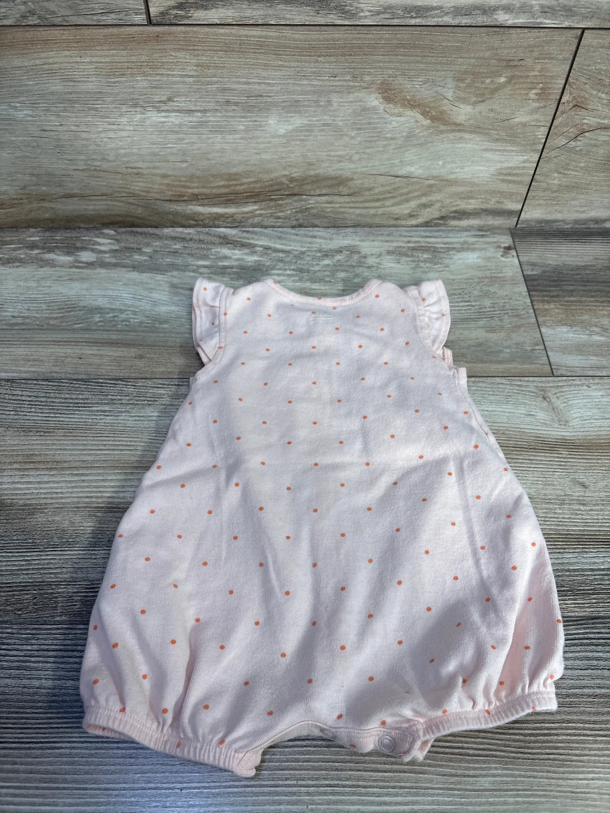 Flat lay of Carter’s peach shortie romper showcasing orange polka dots and elastic leg openings on a wood floor