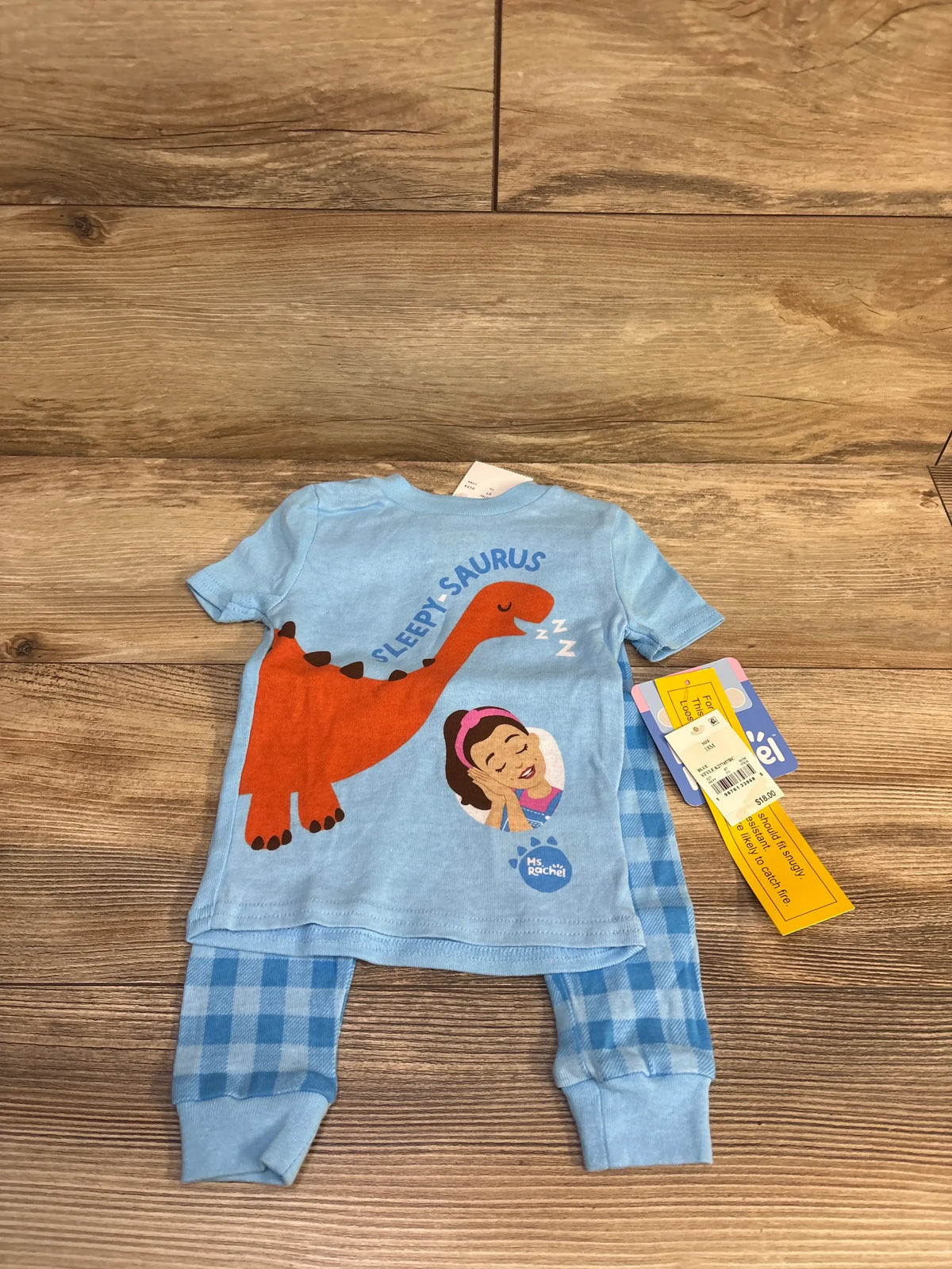 Flat lay of 2-piece Sleepy-Saurus pajama set with blue tee featuring red dinosaur graphic and blue checkered pants on wood