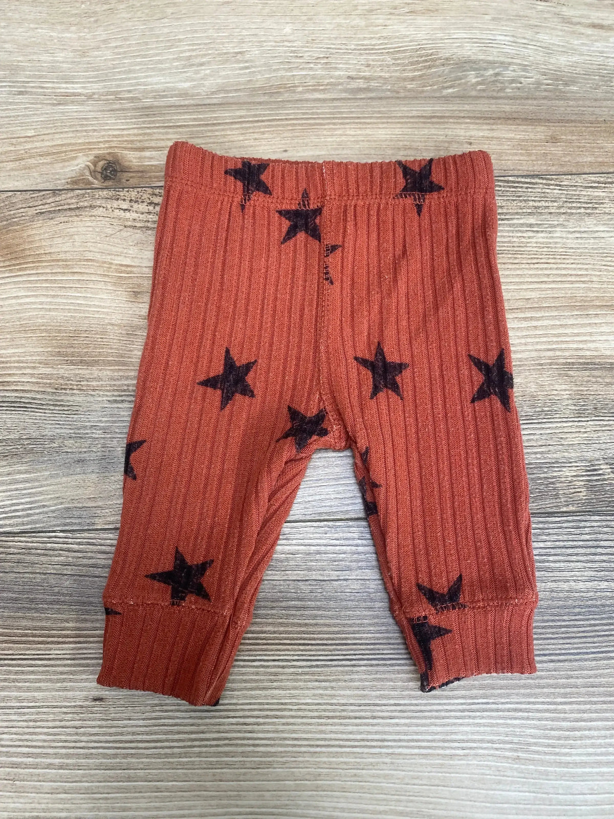 Flat lay of Grayson Collective ribbed newborn pants in rust with black stars, elastic waist and cuffed ankles on light wood