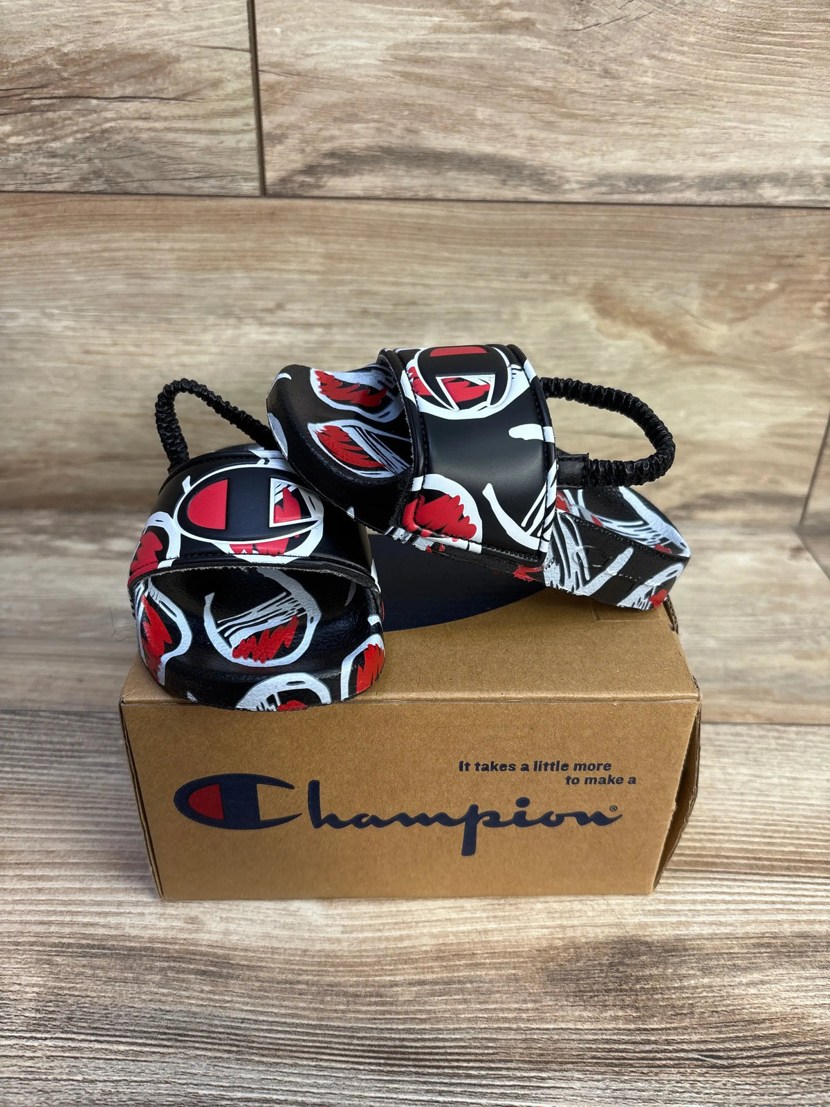 Front view of black Champion Comic slide sandals showcasing red and white comic graphics on a cardboard shoebox