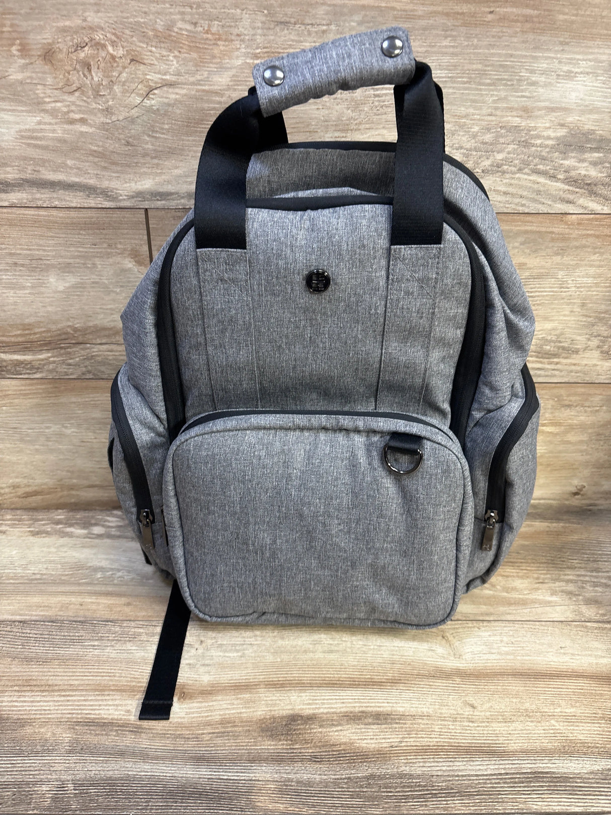 Front view of Huggies Signature Pack diaper bag showcasing gray heather fabric, front zip pocket, side pockets, and padded top handle with wood background